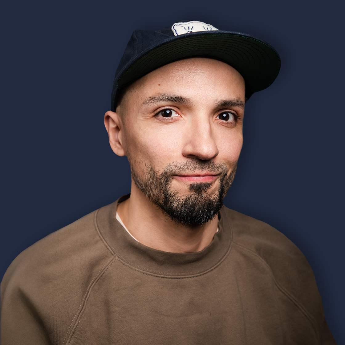Close-up of a young man with a beard, wearing a dark baseball cap and a brown sweatshirt, smiling slightly against a dark blue background.