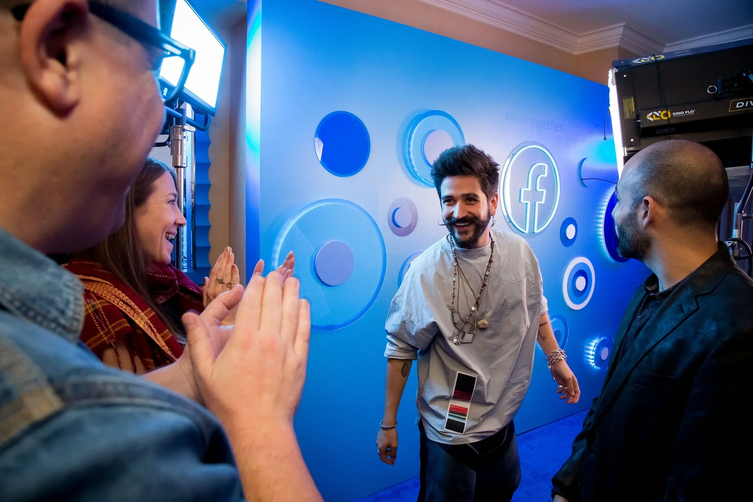 A group of five people standing close together in front of a blue wall with illuminated social media logos. They are smiling, engaging in conversation, and appear to be at a social or professional event.