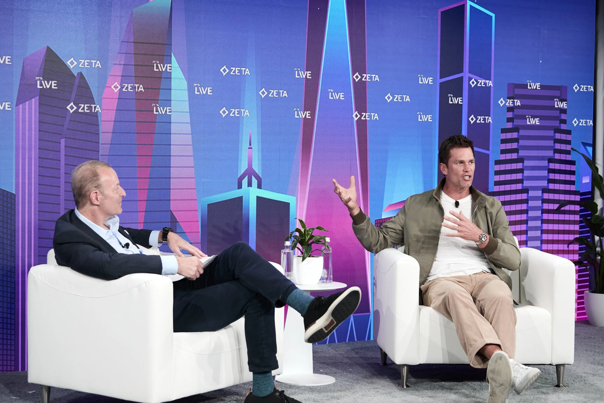 Two men are seated on stage, engaged in a discussion. The man on the right is speaking gesturing with his right hand, wearing a green jacket and beige pants. The man on the left is listening, wearing a dark suit. The background features a colorful cityscape graphic with the words 'ZETA' and 'LIVE' repeated.