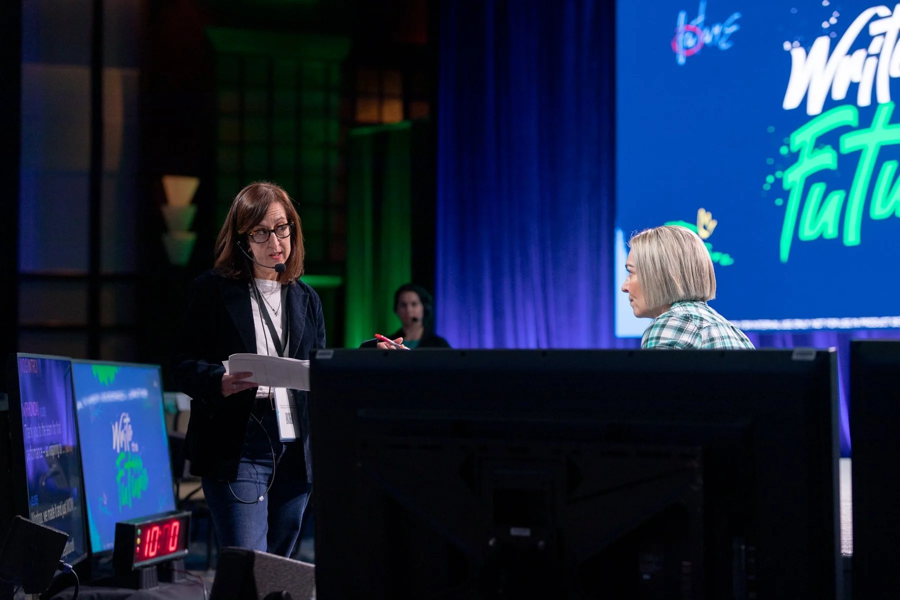 Two women having a conversation at a conference, with monitors showing the event's branding and a large screen in the background displaying 'We Rise To'. The woman on the left is holding papers and talking, while the woman on the right is listening.