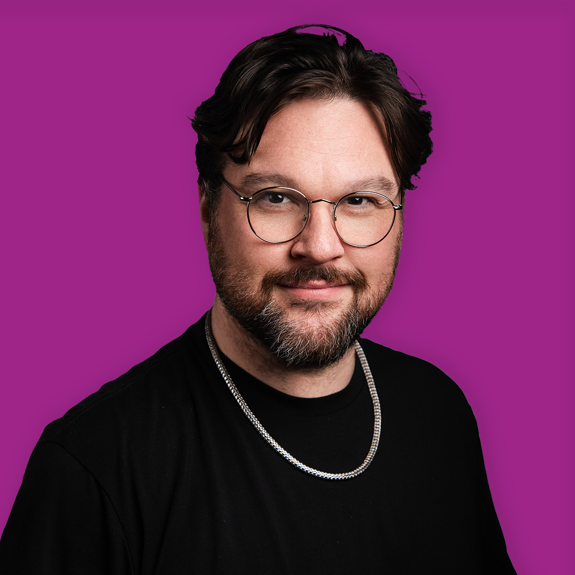 A man with glasses and a beard smiling, wearing a black shirt and a silver chain, against a purple background.