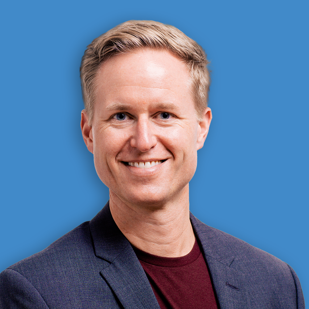 A smiling man with blonde hair wearing a dark blazer and maroon shirt against a blue background.