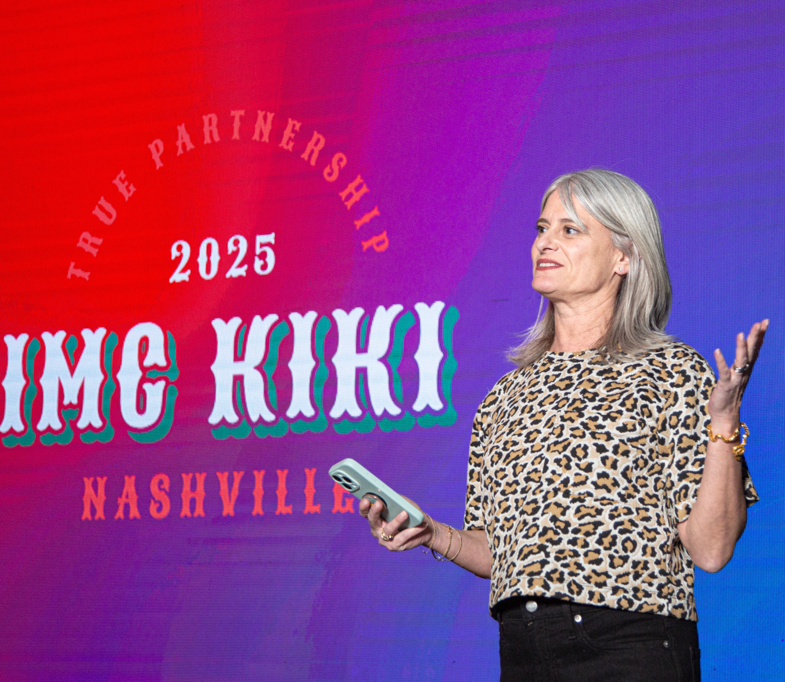 A woman with gray hair wearing a leopard print shirt holding a smartphone and speaking on stage next to a colorful background with text promoting the 2025 event 'Umphrey's Party' in Nashville.