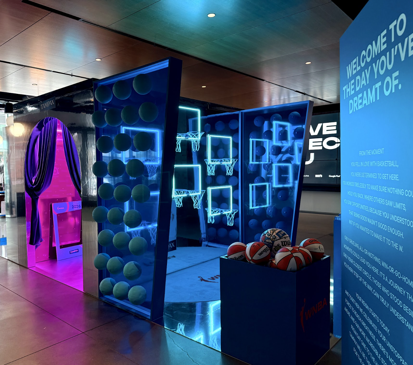A sports-themed indoor display with a blue neon basketball hoop setup enclosed by lit blue frames, a box of Wilson basketballs, and a purple curtain with a pink digital sign area. To the right, a large blue informational panel contains motivational text.