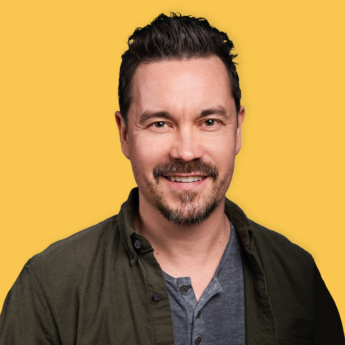 A smiling man with dark hair and a beard, wearing a dark green button-up shirt over a gray t-shirt, posed against a solid yellow background.