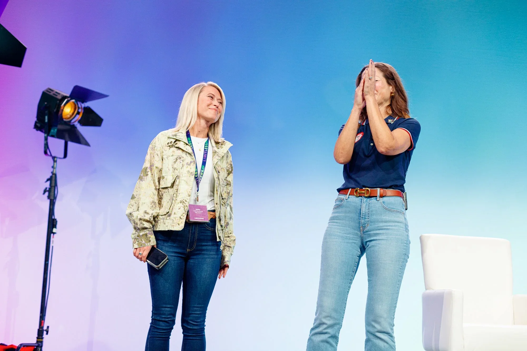 Two women on stage, one smiling at the other, who has her hands over her face in a gesture of gratitude or emotion, with professional lighting and a vibrant background.