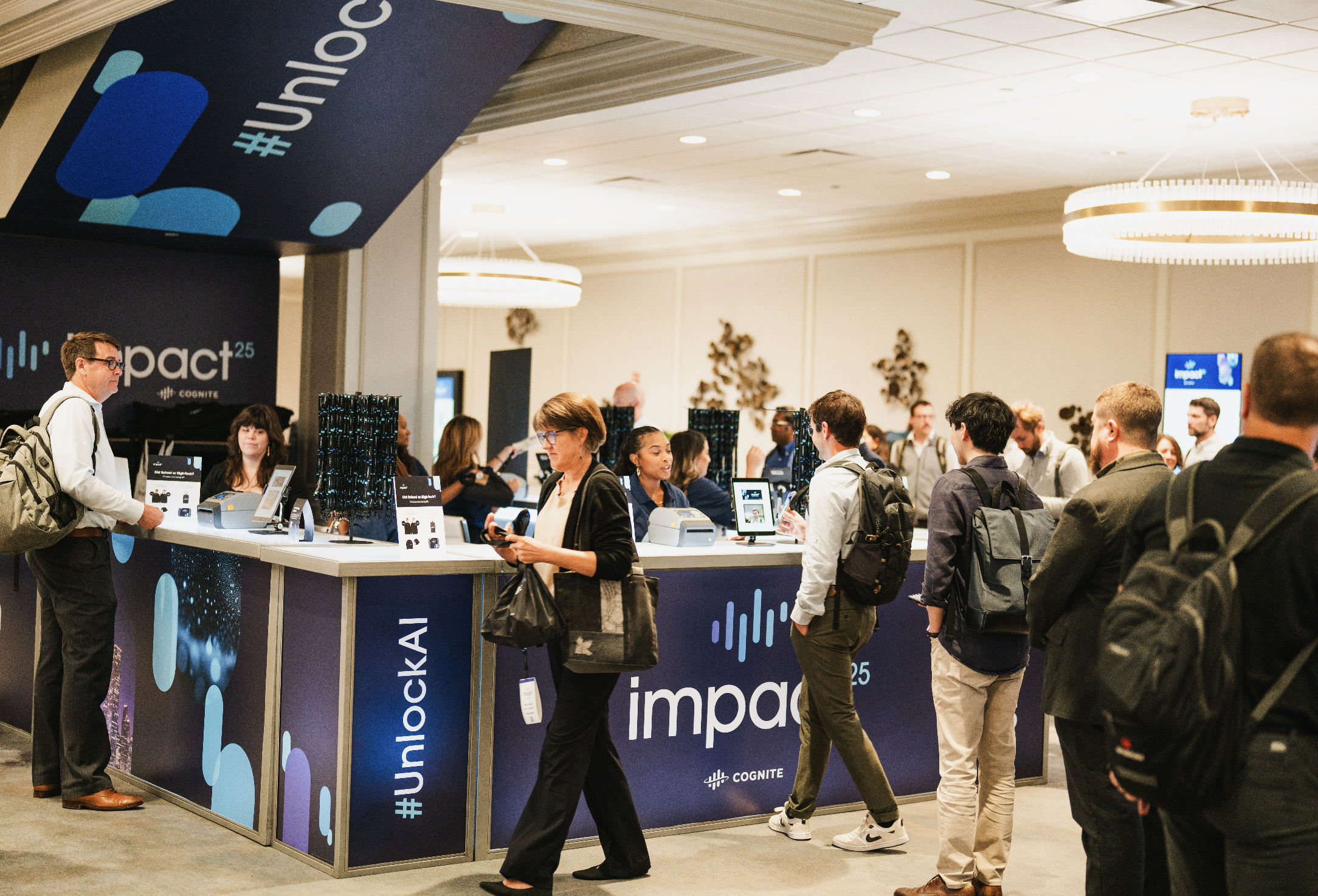 People gathered around an exhibition booth for Impact25 at a conference, with signage displaying the Impact25 logo and social media hashtag.