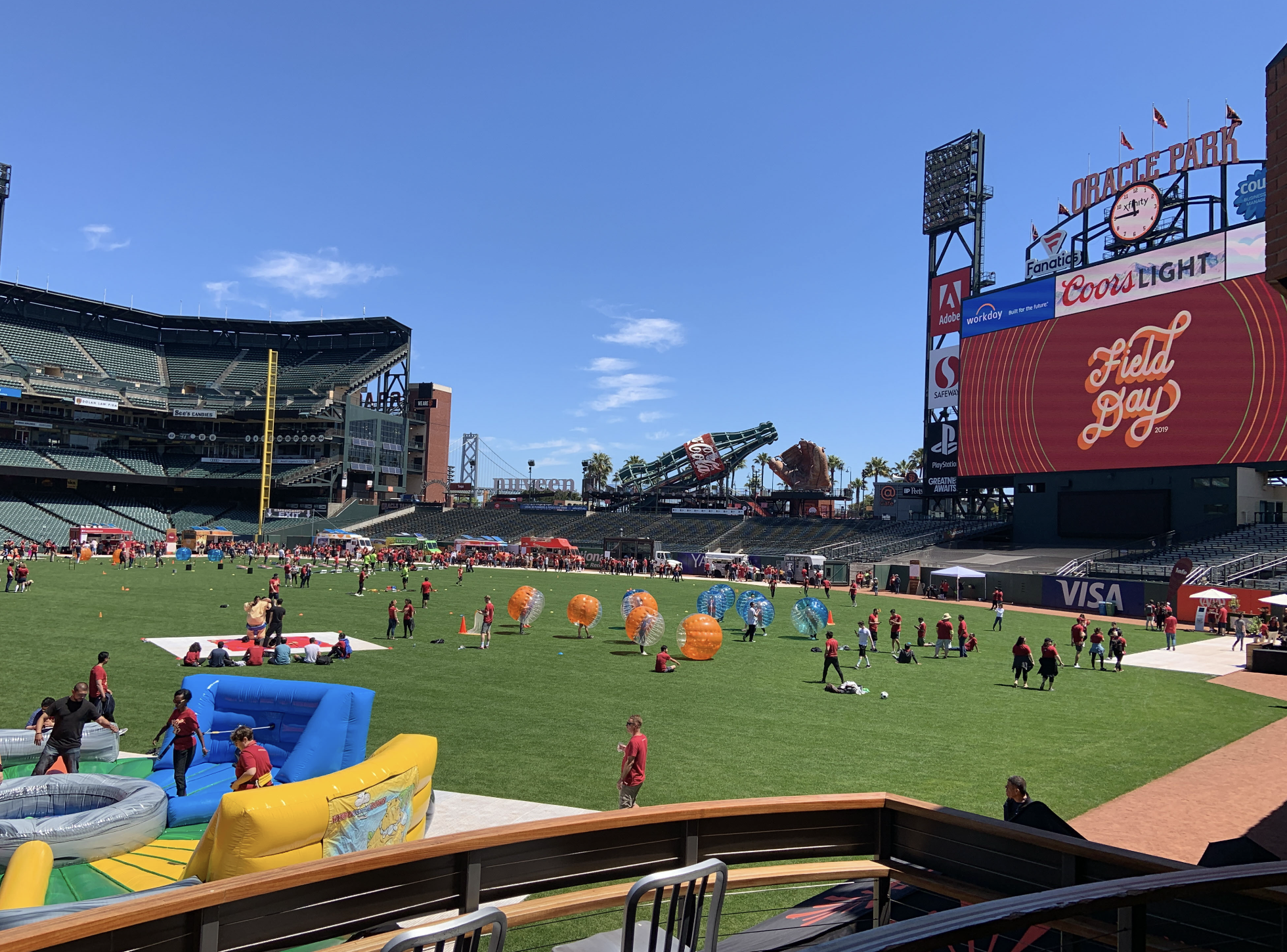 View of a baseball stadium with a large digital screen displaying an event called 'Field Day 2019'. The field is filled with children and adults participating in activities, including inflatable bounce houses and bubble soccer. The stands are mostly empty, and the sky is clear with few clouds.