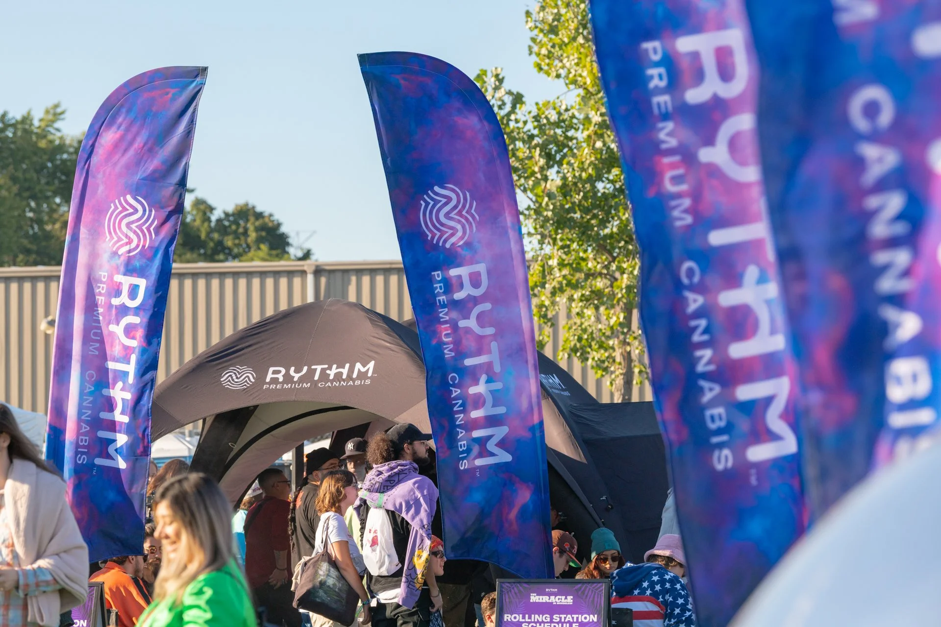 Blue and purple flags with the word 'RYTHM' and 'PREMIUM CANNABIS' printed on them, billowing in the wind at an outdoor event with a crowd of people and a booth labeled 'ROLLING STATION' in the background.