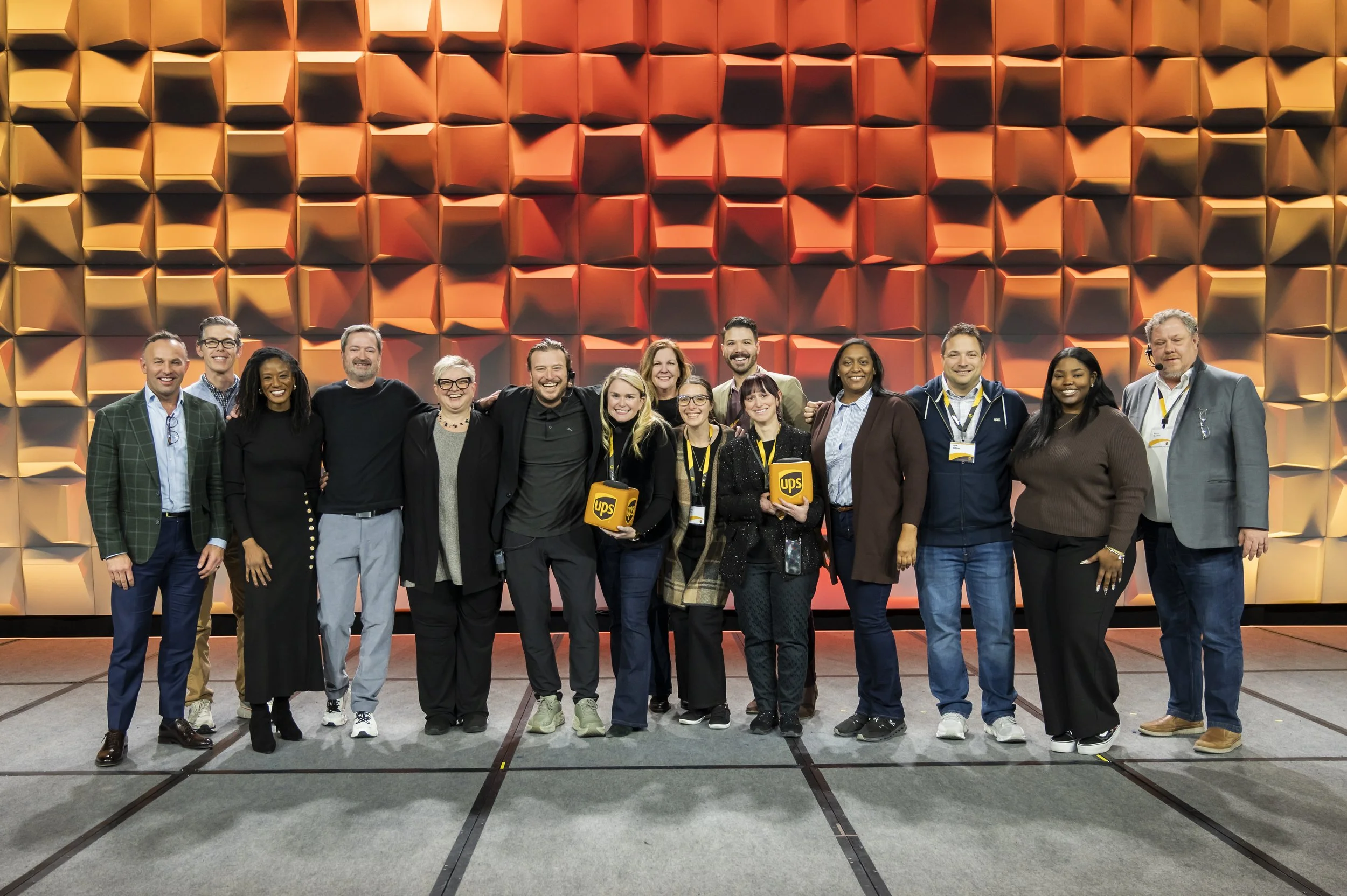 Group of 14 diverse people standing together on a stage with an orange and yellow geometric backdrop, smiling and posing for a photo.