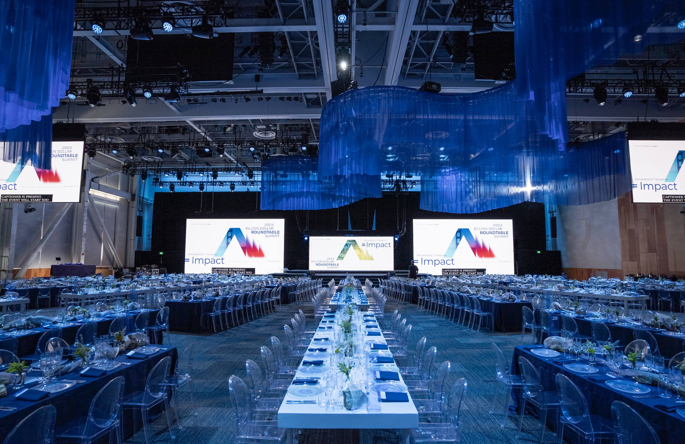 A large conference or banquet hall set up for an event with long tables arranged for numerous attendees, decorated with small flower arrangements, and multiple large screens displaying the event's branding and themes, including '2024 Billion Dollar Roundtable Summit' and 'Impact'.