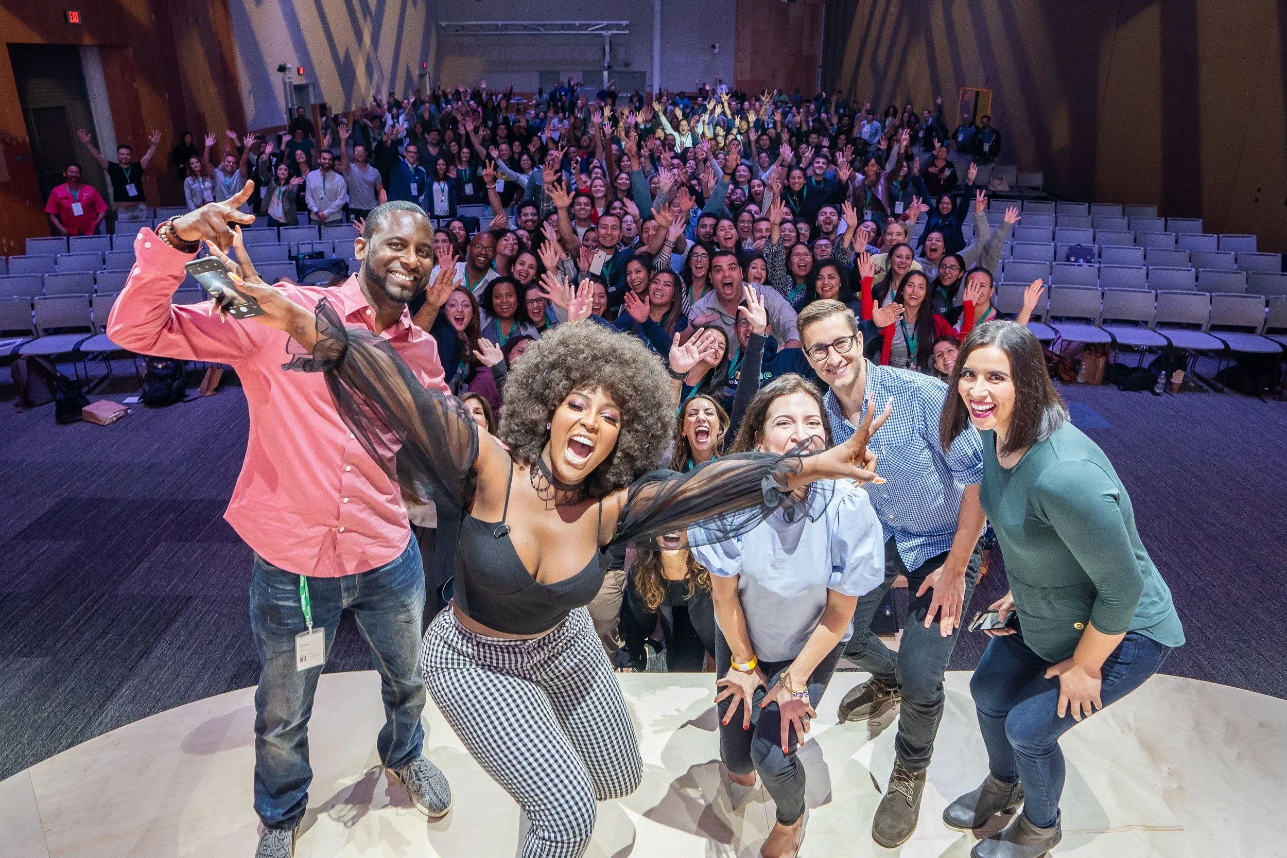 Group selfie of diverse smiling people at a conference or event, waving and looking happy in an auditorium.