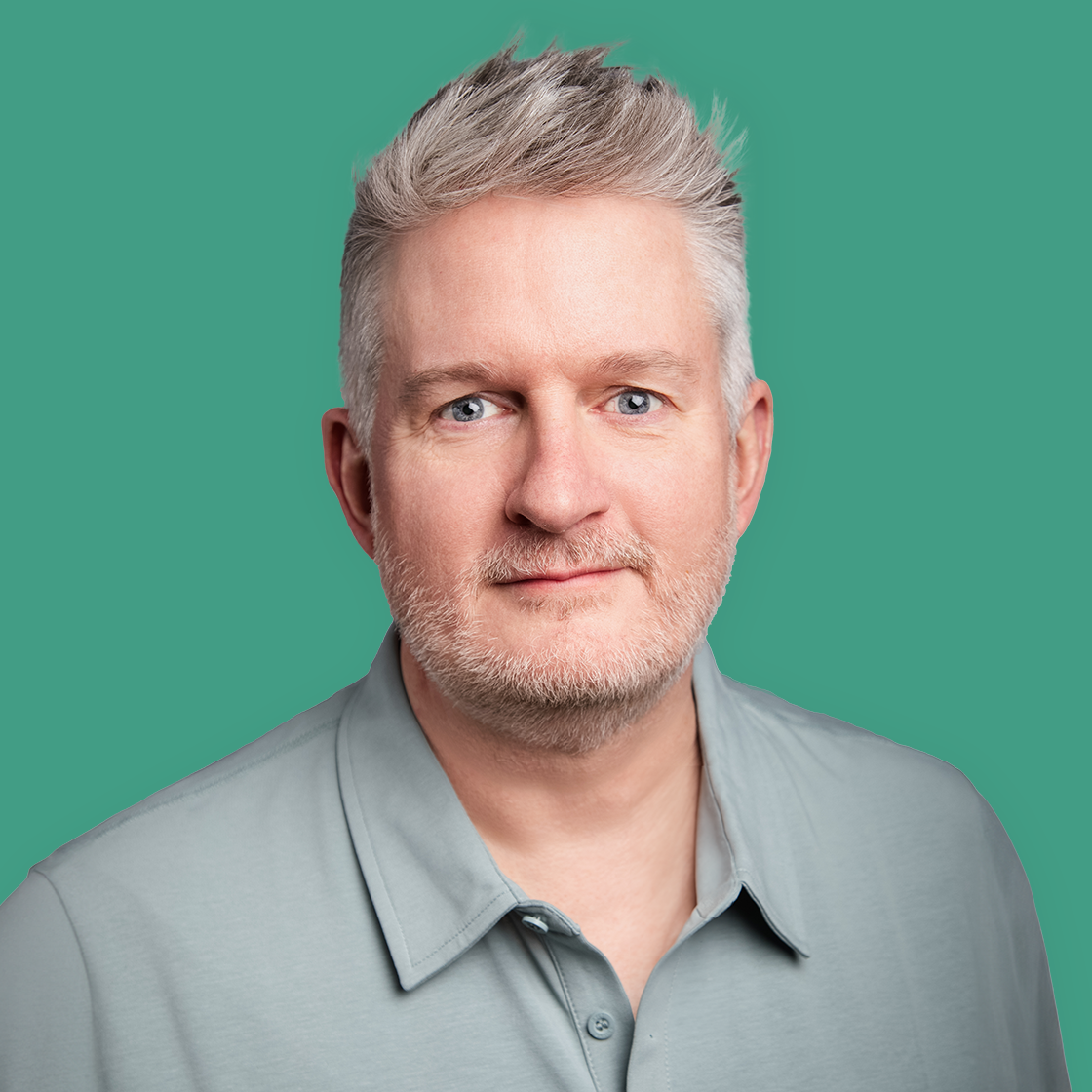 A middle-aged man with gray hair and beard, wearing a gray collared shirt, looking at the camera with a neutral expression against a teal background.