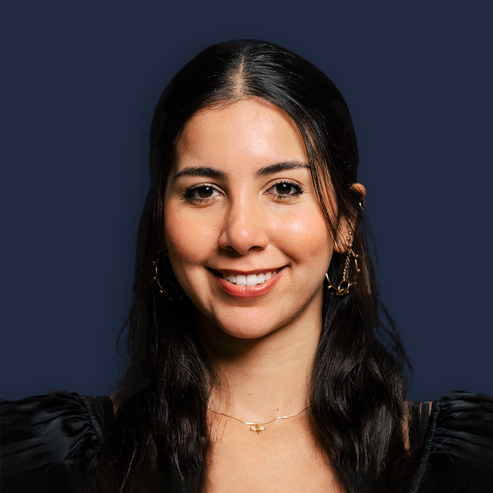 Close-up photo of a young woman with dark hair, smiling, wearing earrings and a delicate necklace, against a dark background.