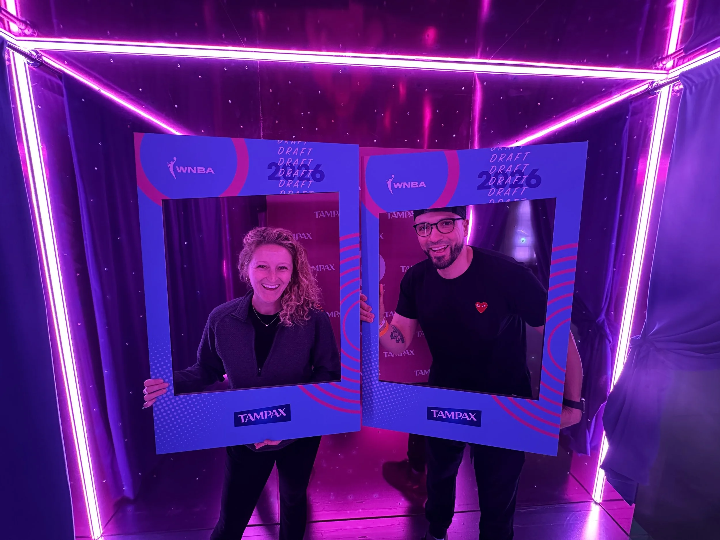 Two people smiling and holding photo frames with the Tampax logo inside a neon-lit, pink and purple room.