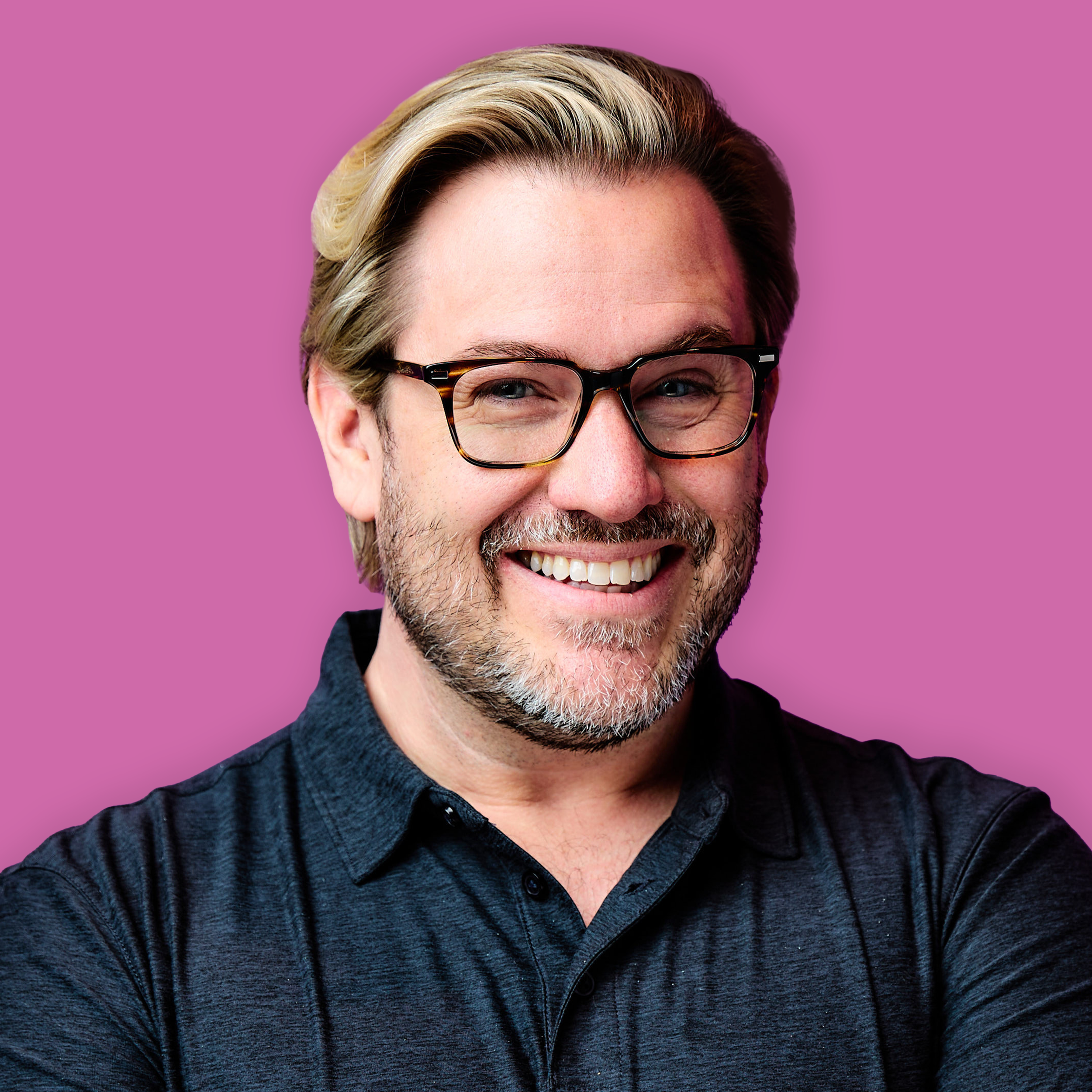 Close-up of a smiling man with gray hair, wearing glasses and a dark blue shirt, against a purple background.