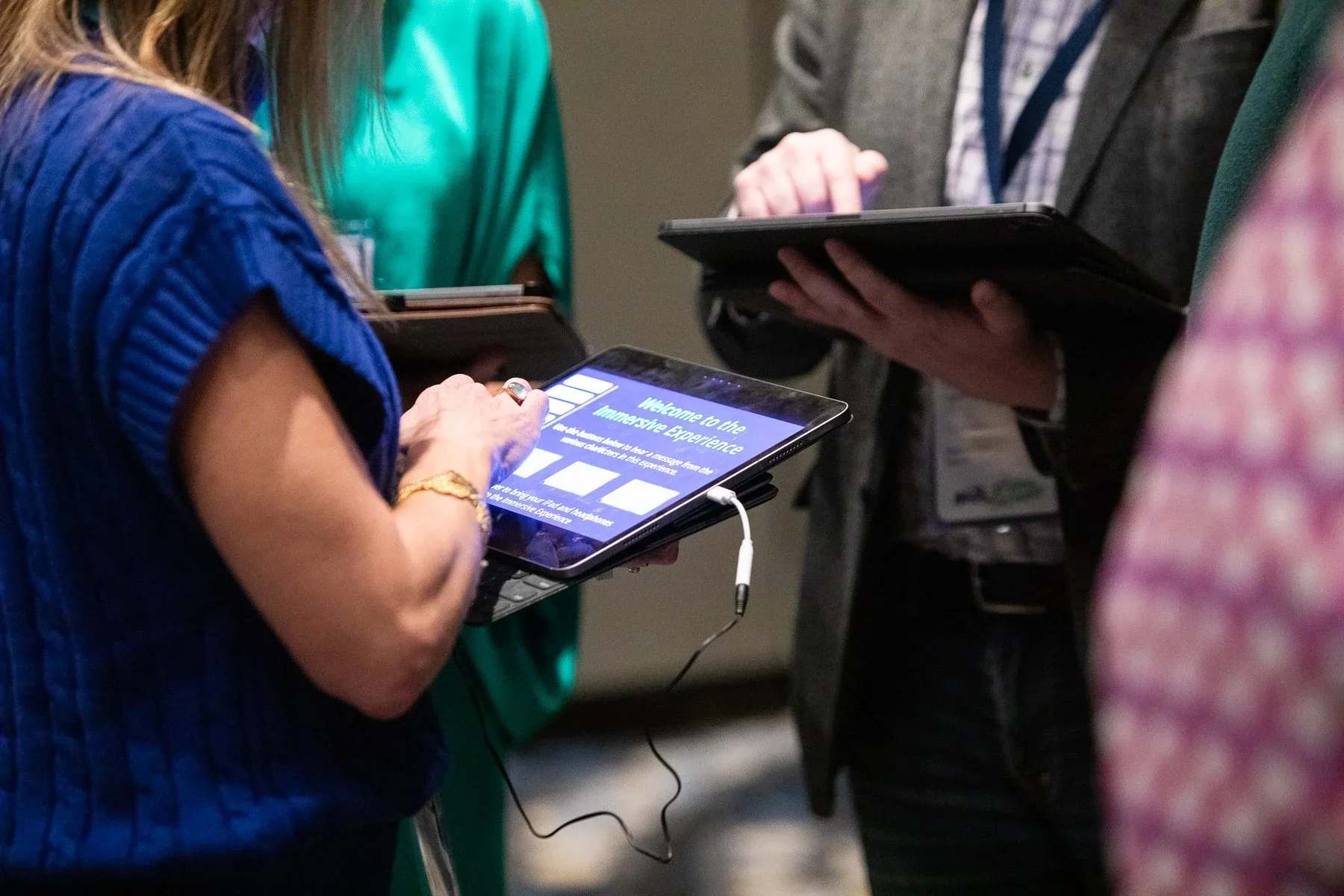 People gathered at a conference or event, looking at and interacting with digital devices, including tablets displaying a welcome message for an immersive experience.
