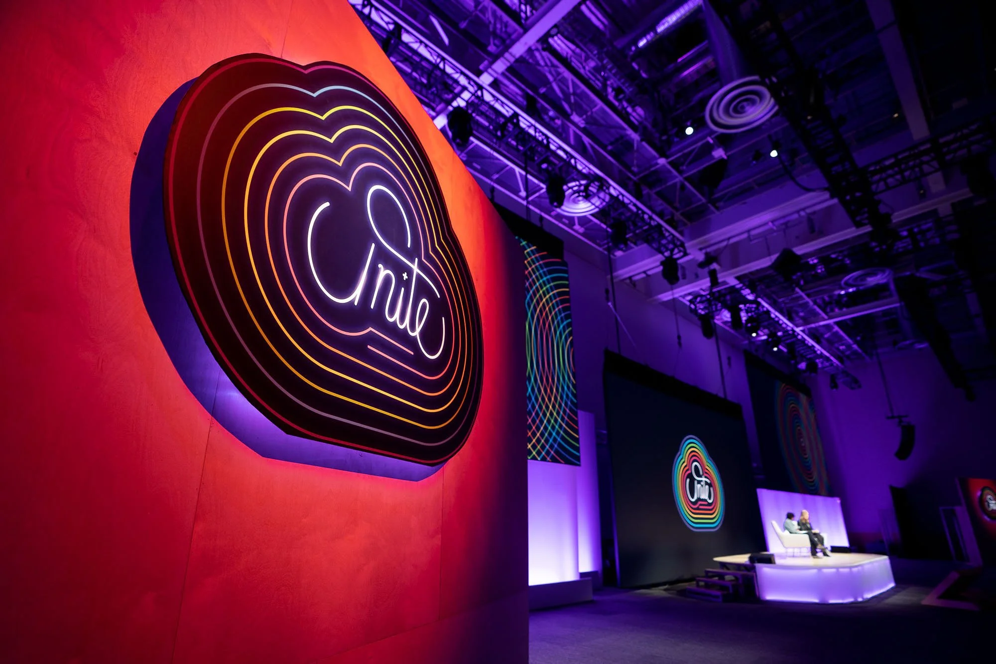 Neon sign with a heart and the word 'unity' inside, displayed on a red wall at a vibrant event space with purple lighting and a high ceiling.