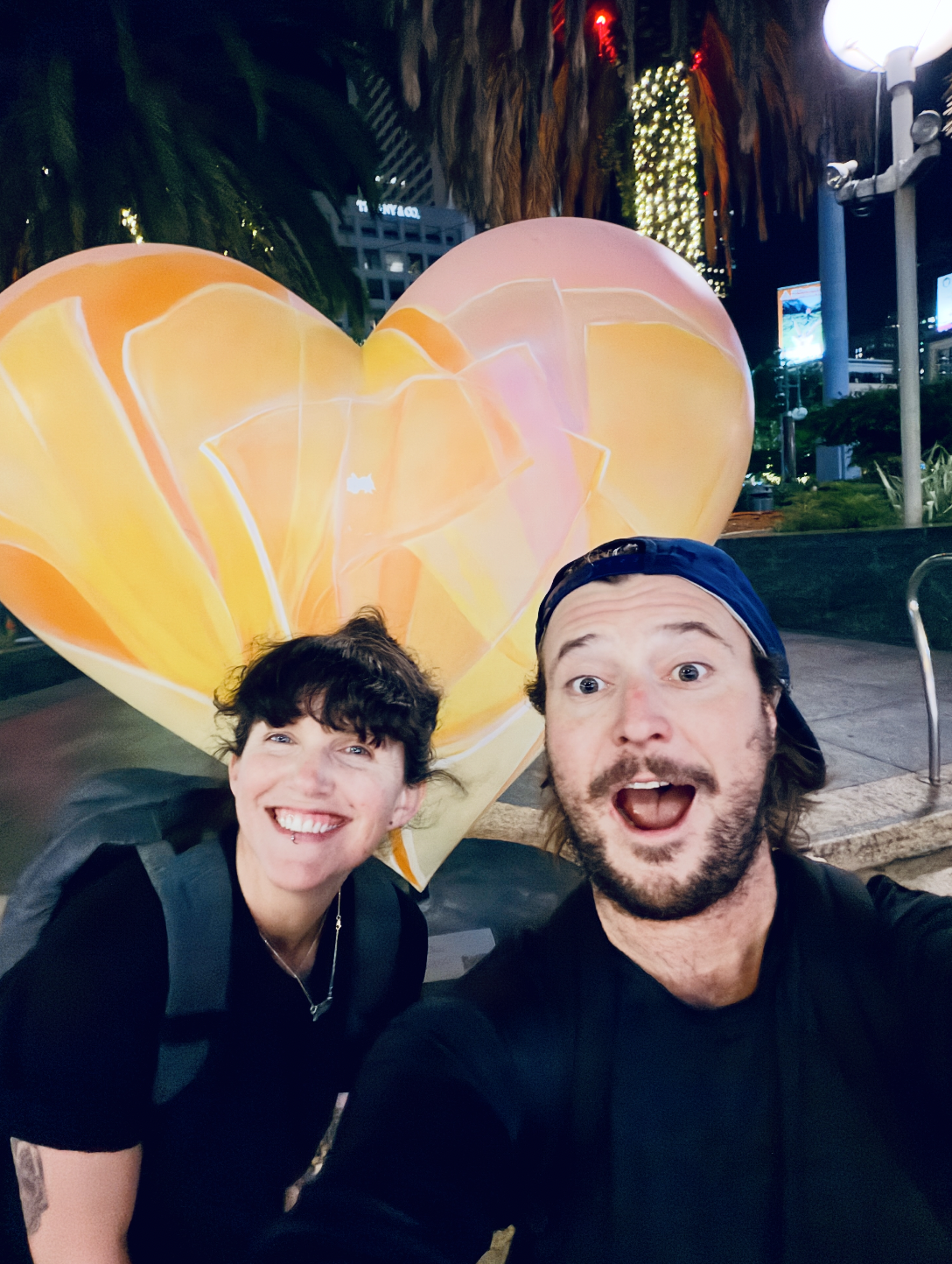A man and woman taking a selfie at night in front of a large, illuminated heart-shaped sculpture with orange and pink hues, outdoors with palm trees and city buildings in the background.
