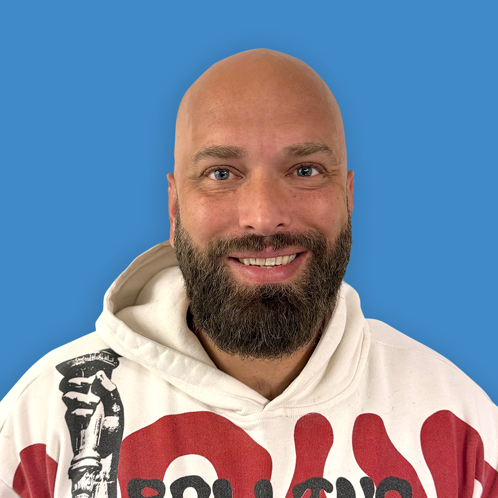 A smiling man with a bald head and a full beard, wearing a white hoodie with red and black graphics, standing against a solid blue background.