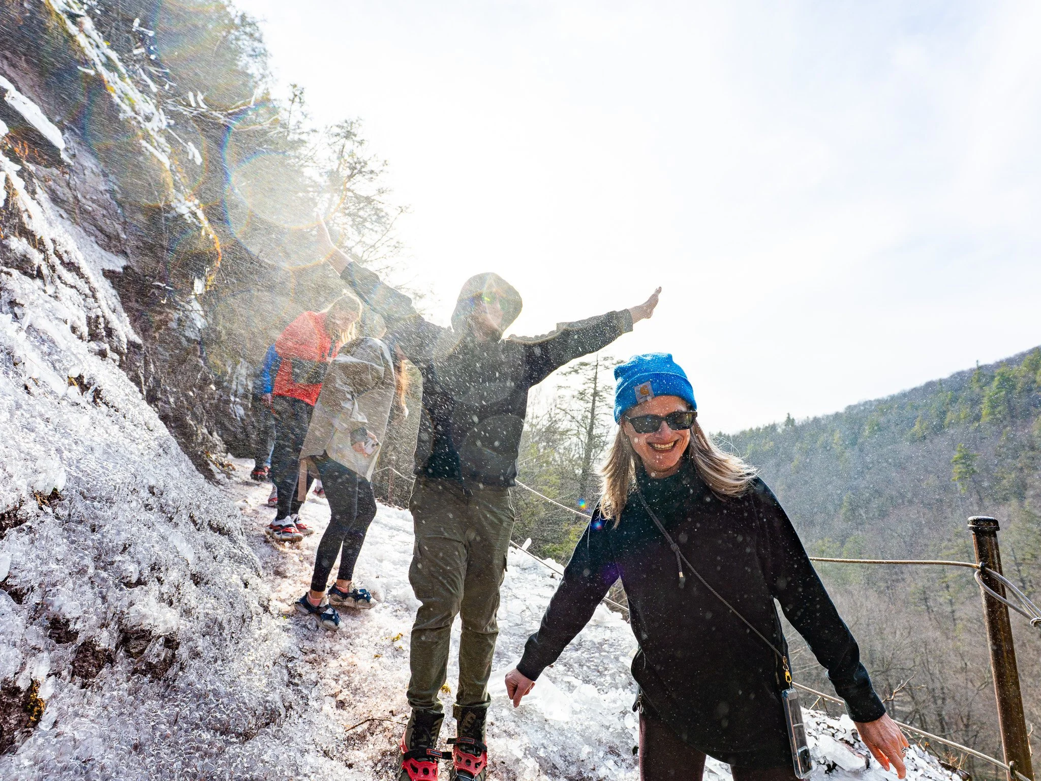 Group of people hiking on a snowy trail in a forested mountainous area, with some wearing sunglasses and winter gear, and a woman smiling in the foreground.
