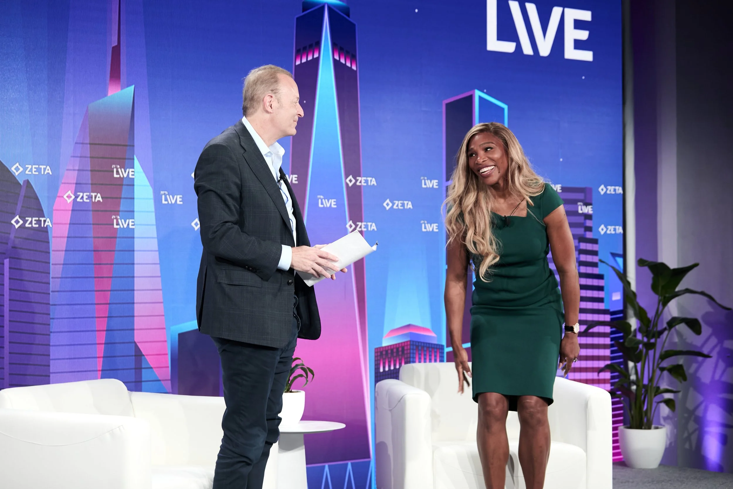 A man and a woman are on a stage with a blue backdrop that has city buildings and the text 'ZETA LIVE'. The man is wearing a dark blazer and white shirt, holding a paper, and the woman is wearing a fitted green dress, smiling.