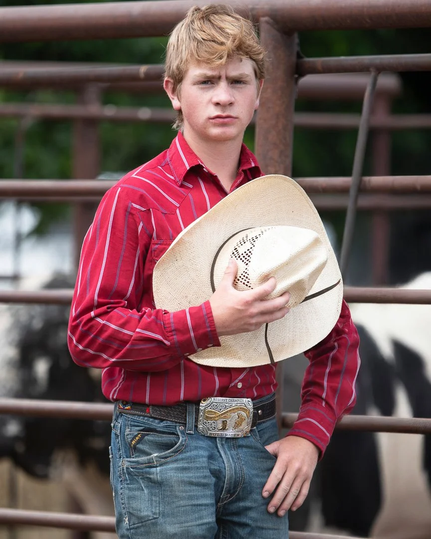 A young man with blonde hair wearing a red striped shirt and blue jeans, holding a cowboy hat, standing in front of a rustic fence with cows in the background.