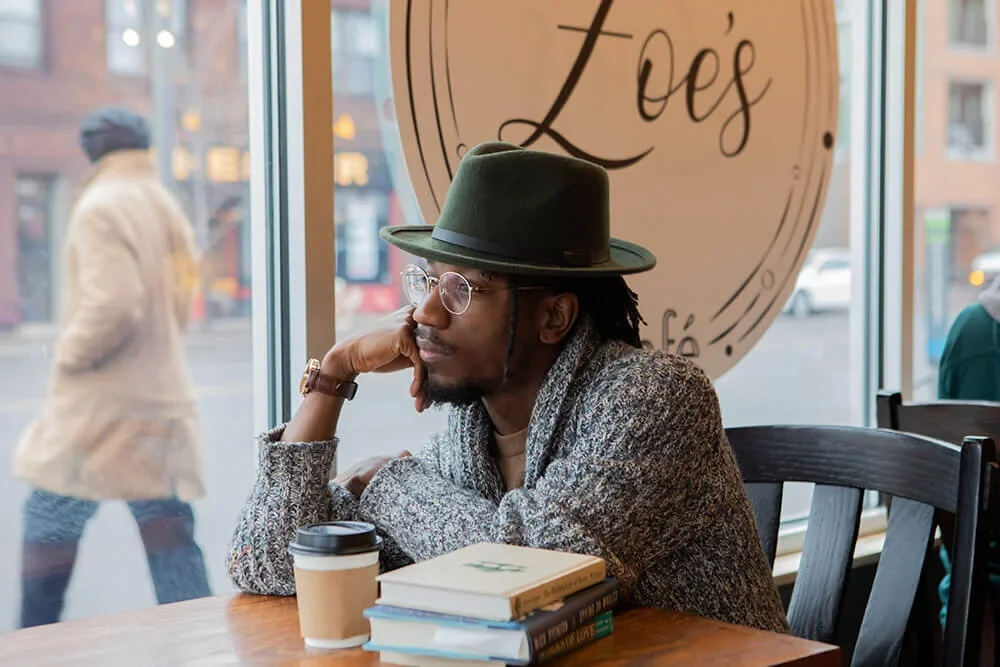A man with glasses and a hat sitting at a café table, looking out the window with books and a coffee cup in front of him.