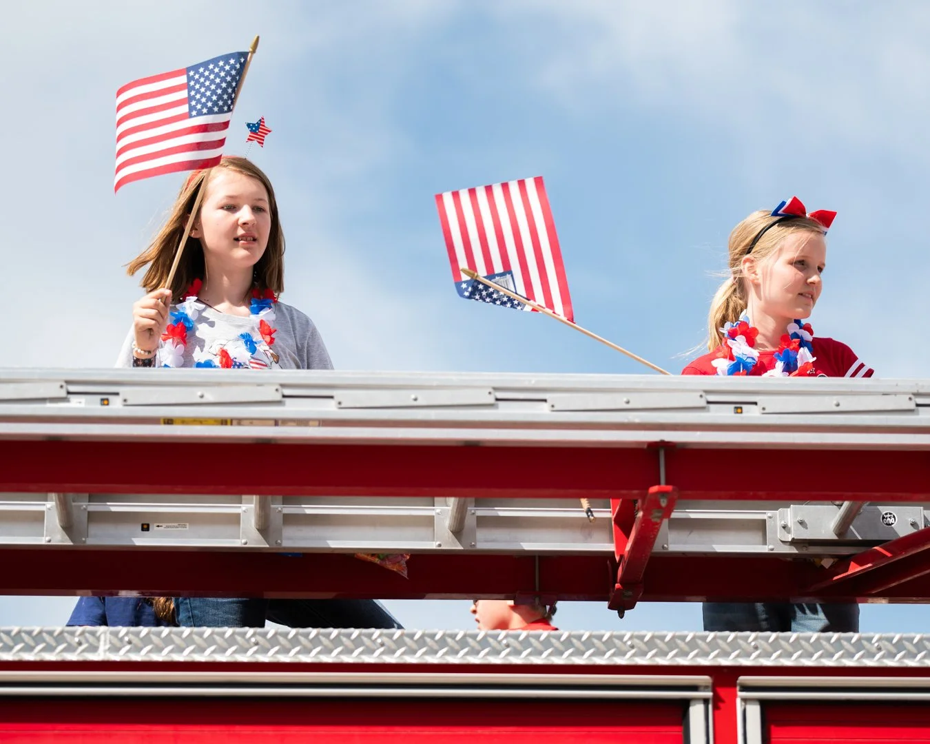 Two young girls with American flags on their shirts and leis, standing on a parade float with a clear blue sky background.