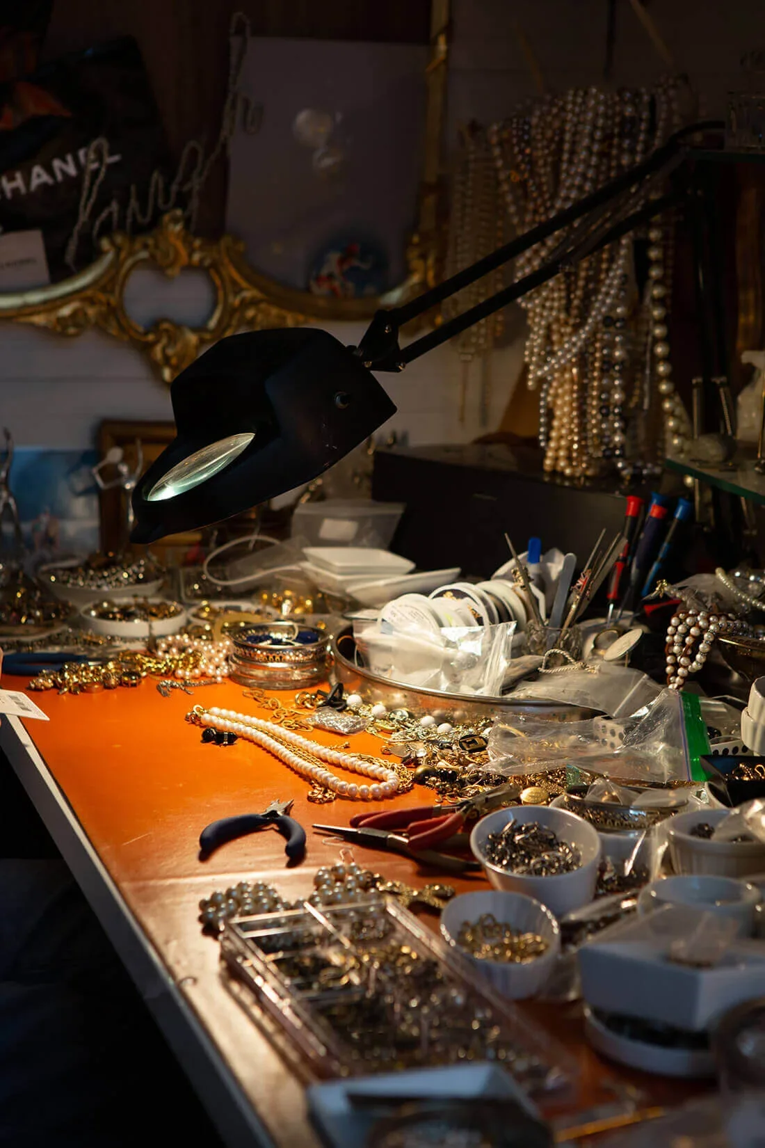A cluttered jewelry workspace with various jewelry pieces, tools, and accessories spread across a wooden table, illuminated by a desk lamp.