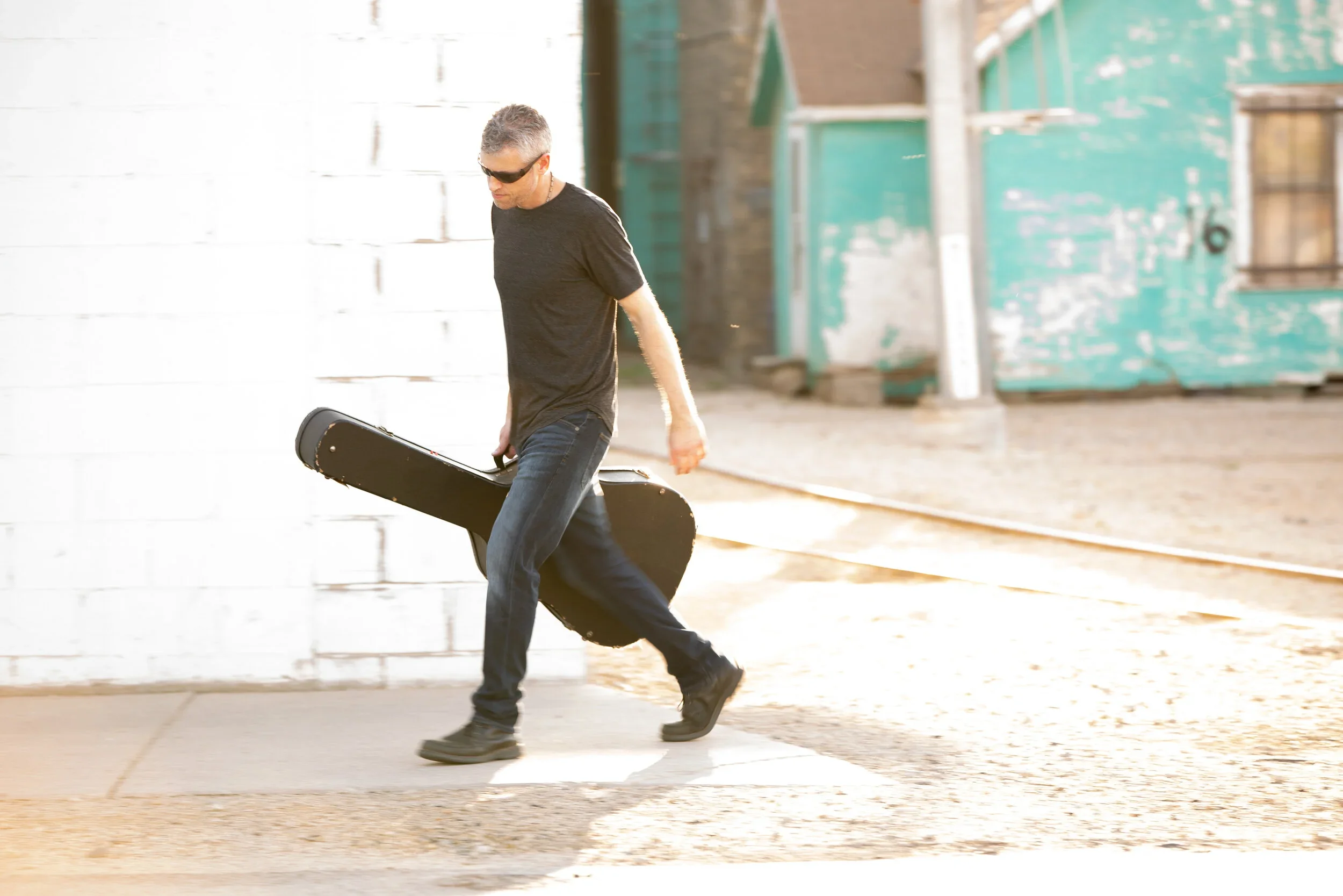 A man dressed in black walking on a sidewalk carrying a guitar case as he passes by an old building with weathered blue-green paint.