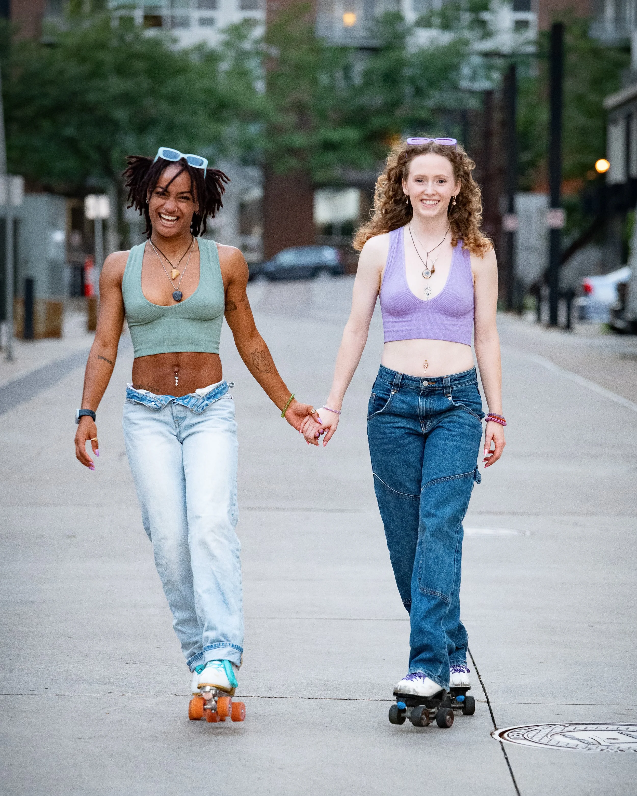 Two young women roller skating hand in hand on a city sidewalk, smiling and wearing casual tank tops and jeans, with sunglasses on their heads and necklaces.