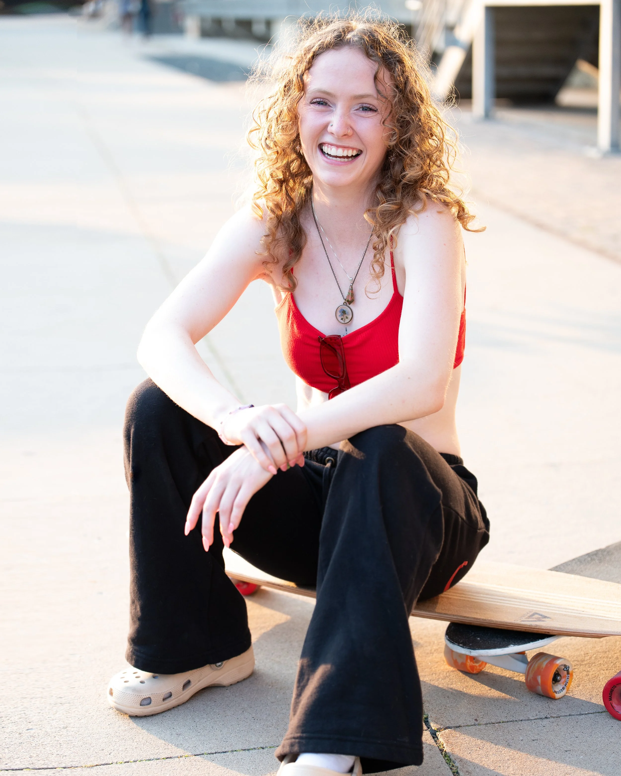 A young woman with curly red hair, wearing a red tank top, black pants, and beige Crocs, smiling and sitting on the ground next to a skateboard at a skate park.