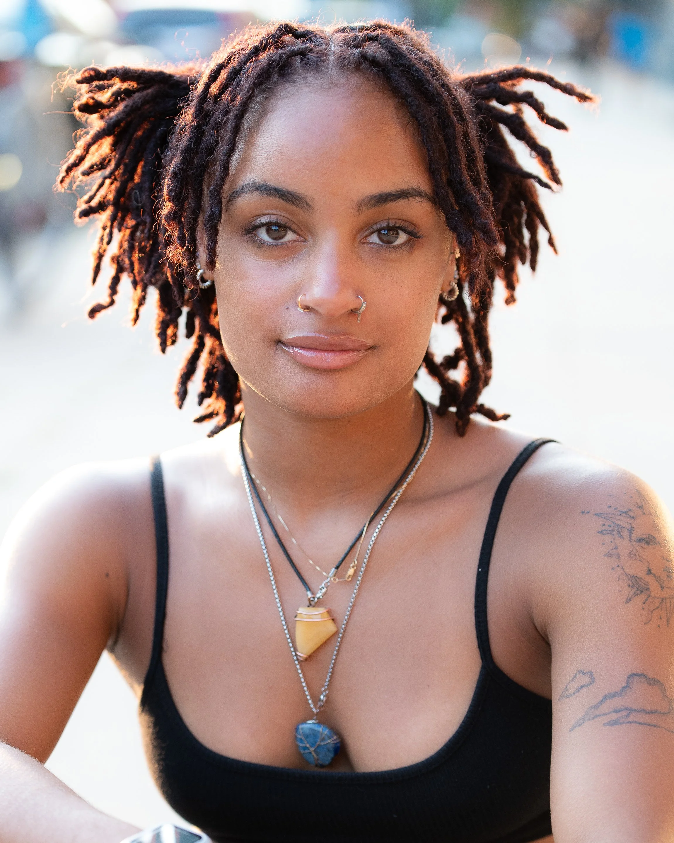 Close-up of a young woman with dreadlocks, wearing a black tank top, multiple necklaces, earrings, and nose piercings, outdoors with a blurred background.