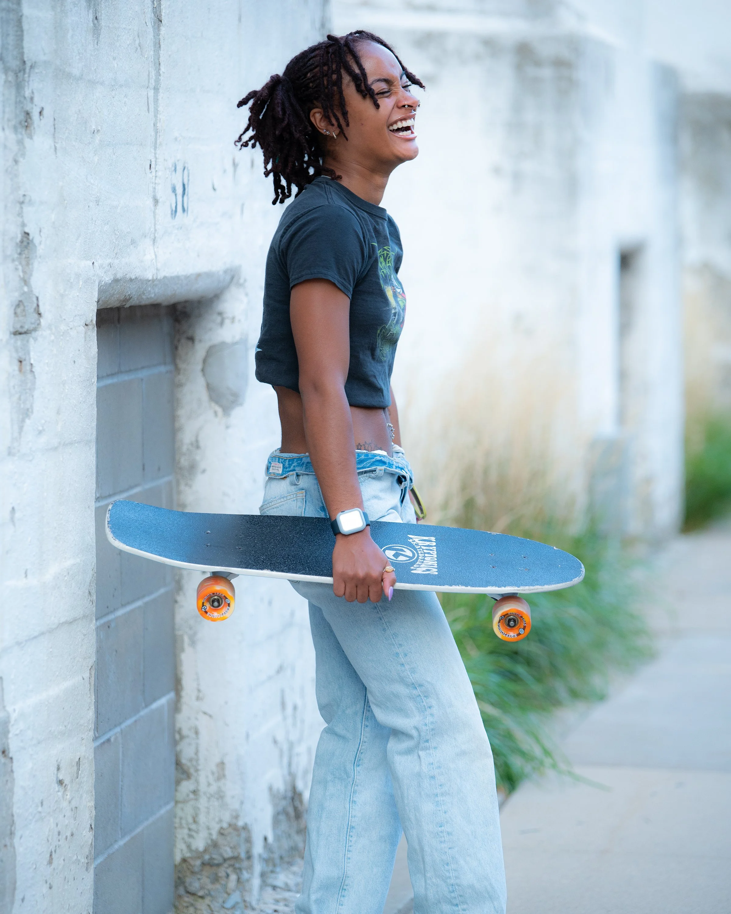 A woman with dreadlocks holding a skateboard, standing against a white brick wall, smiling and laughing.