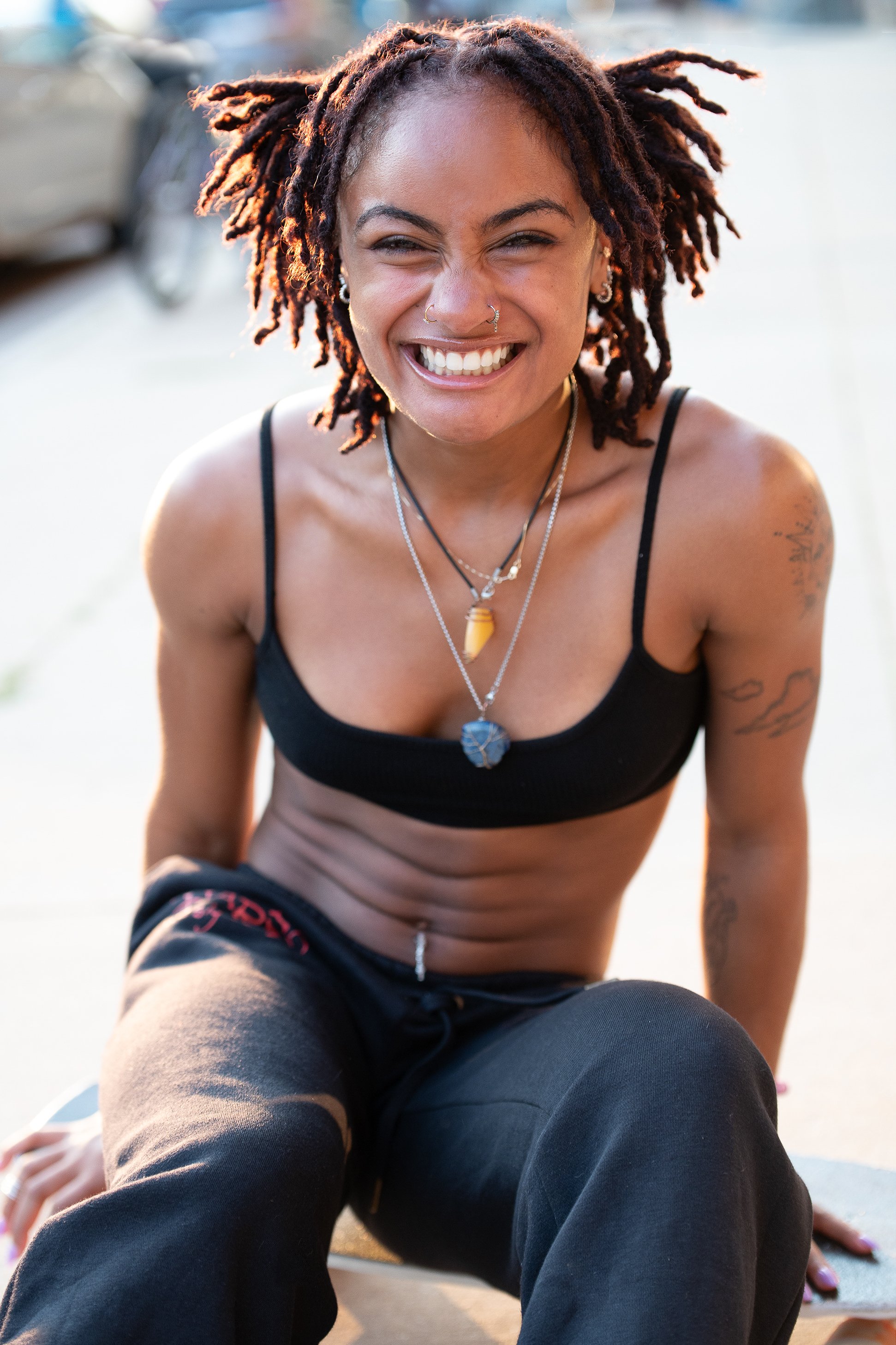 A young woman with dark skin and dreadlocks smiling brightly during an outdoor workout or yoga session, wearing a black sports bra and black pants, with layered necklaces and visible tattoos on her left arm.