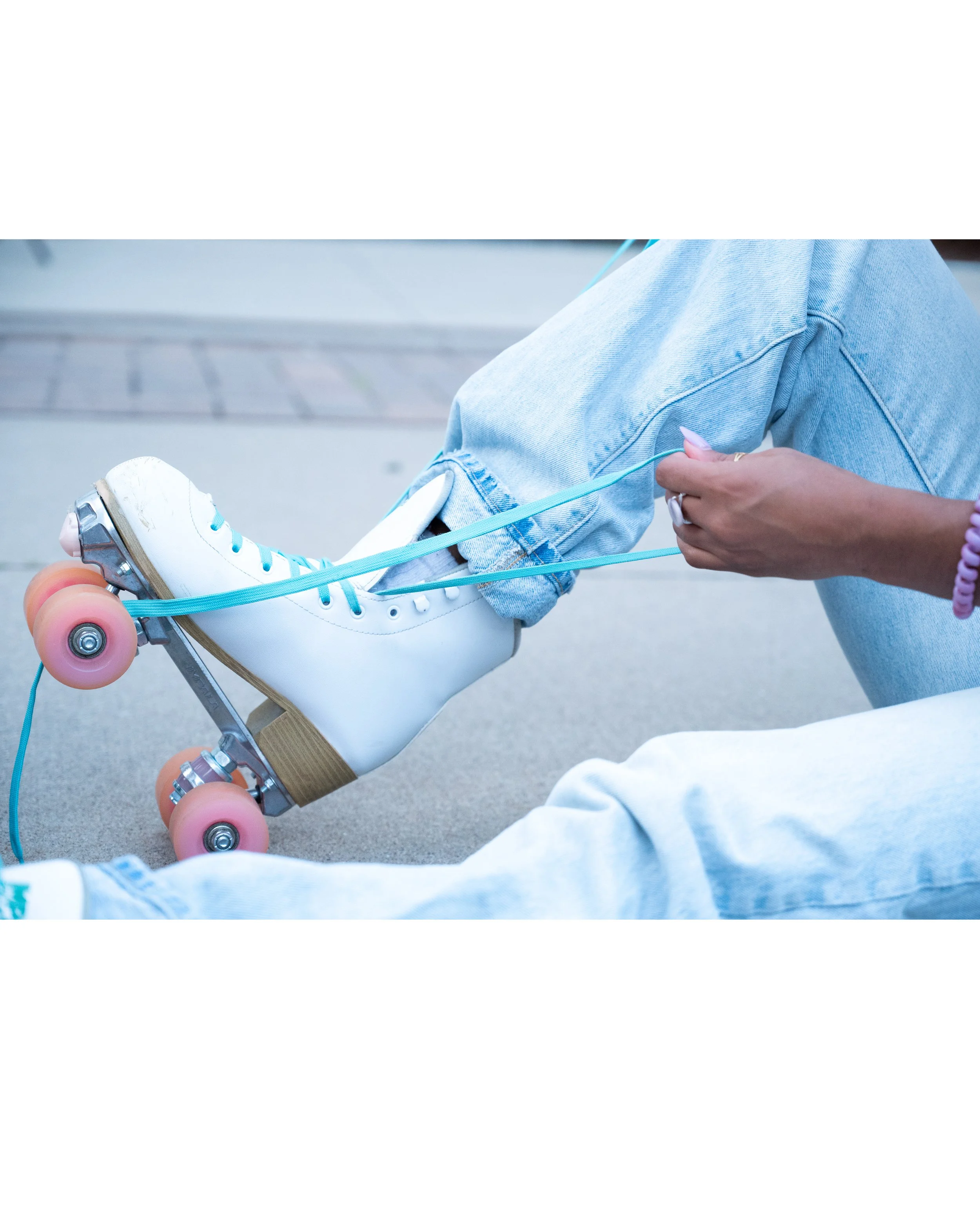 A person sitting on the ground tying the shoelaces of their white roller skates, wearing light blue jeans.