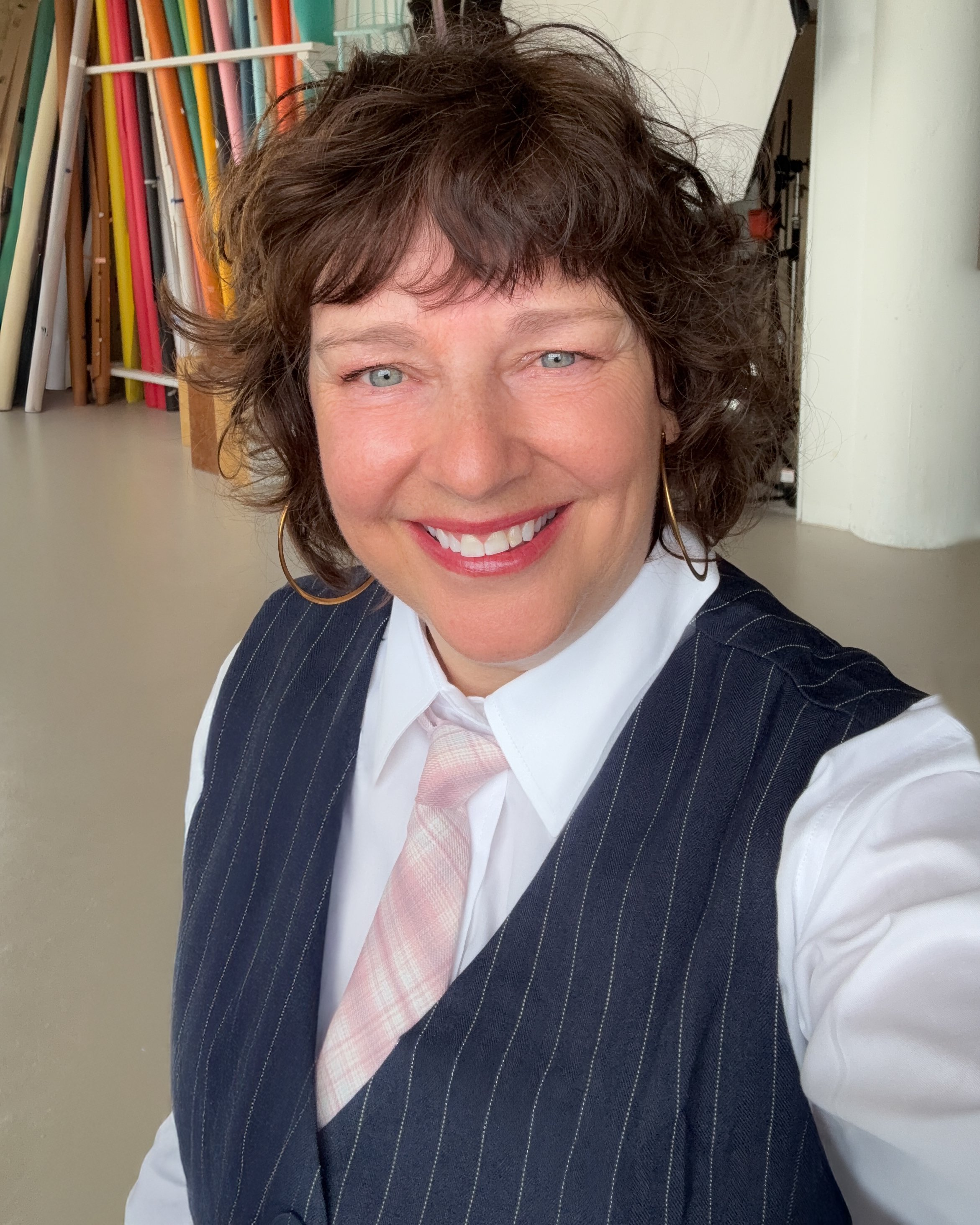 A woman with short, curly brown hair smiling at the camera, wearing a white shirt, a pink tie, and a dark blazer with pinstripes, against a background of colorful rolled papers or materials on shelves.