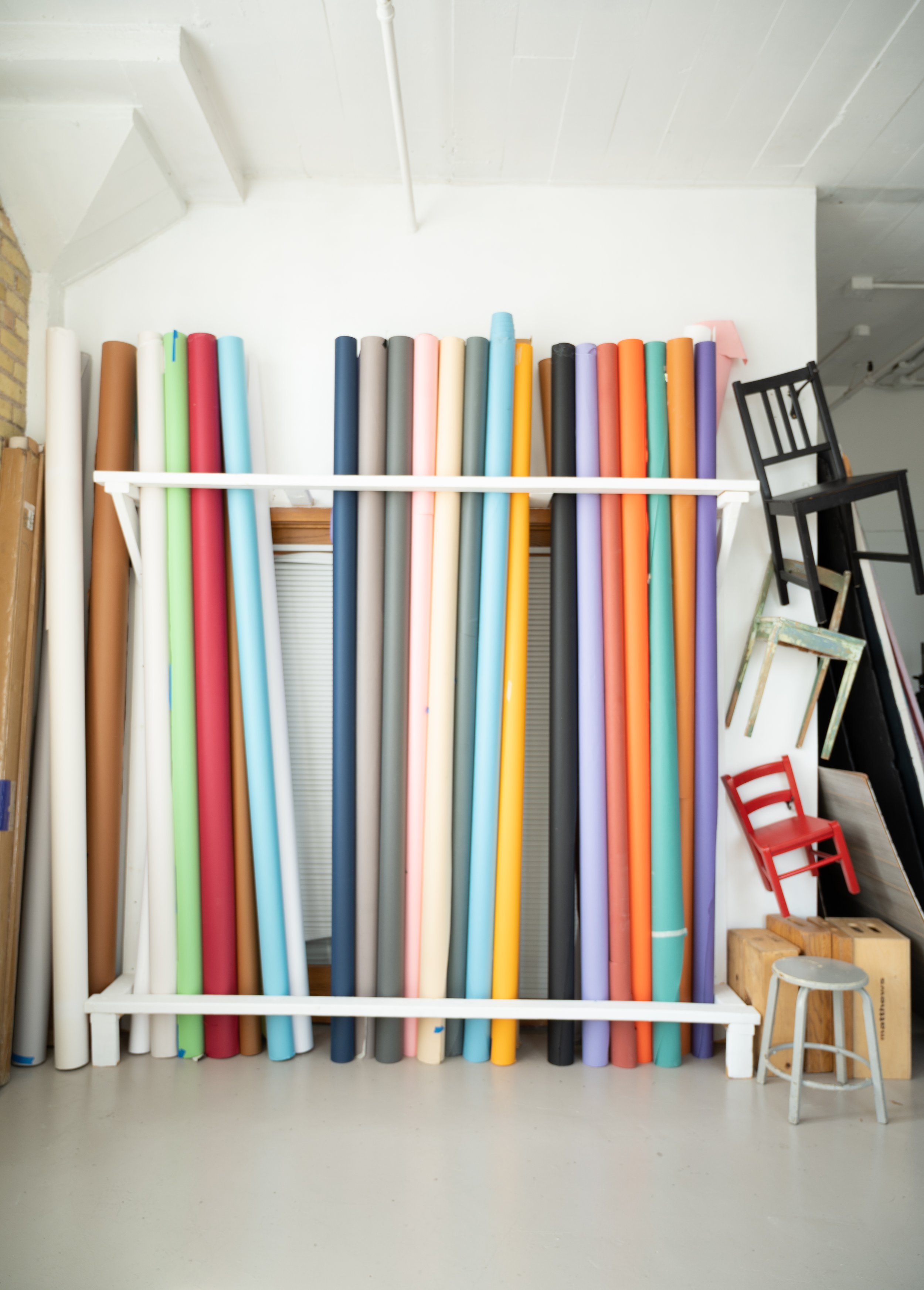 A collection of colorful fabric rolls arranged upright on a white frame, with a black chair, small stools, and a stool with a patterned cushion in a gallery or creative space.