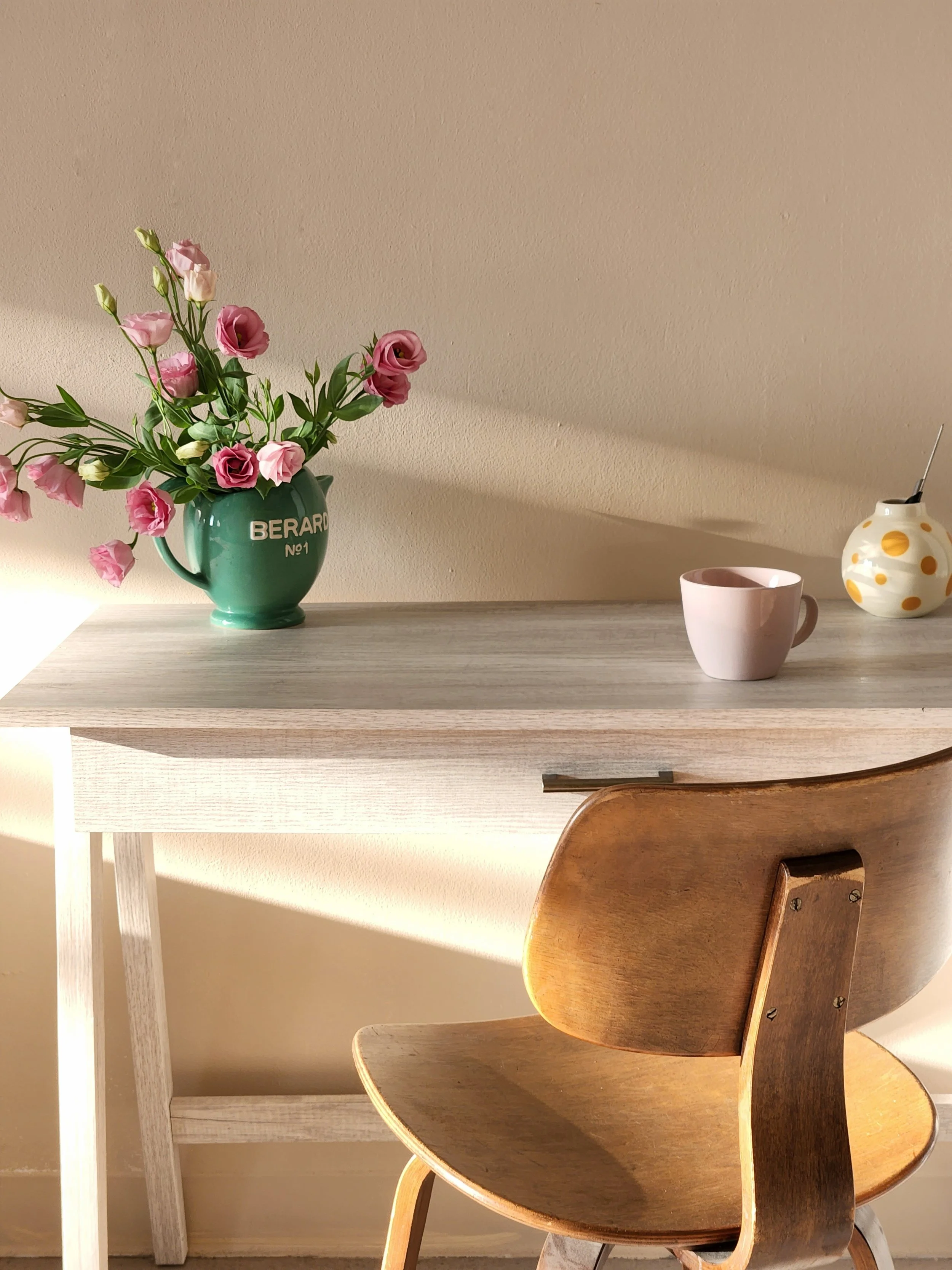 A wooden chair in front of a light-colored wall with sunlight casting shadows. A white table with a potted plant and a small vase on it.