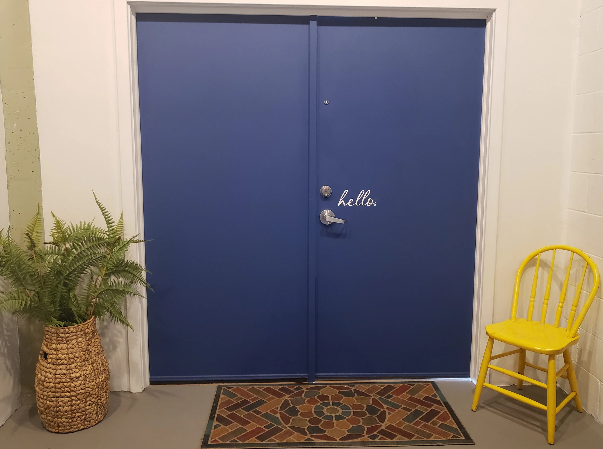 Blue double door with small round peephole and a lock, surrounded by white walls. To the left, a potted green fern plant in a woven basket. To the right, a yellow chair. In front of the door, there is a decorative floor mat with a geometric pattern.