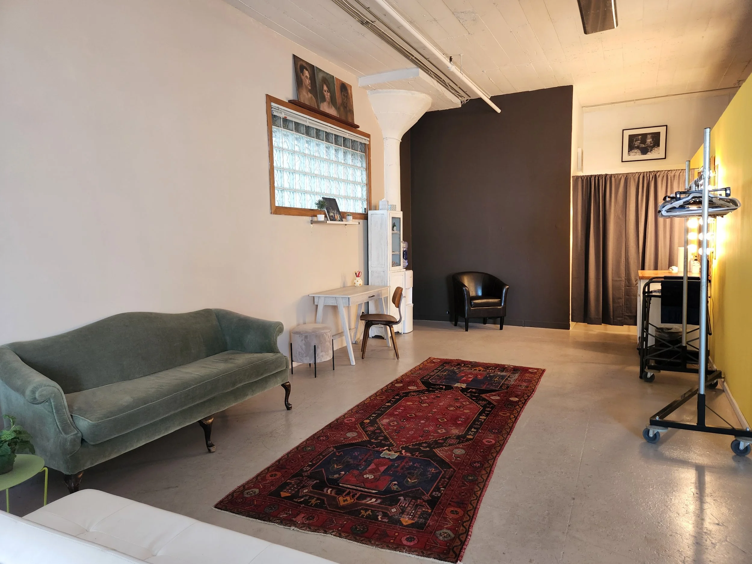 Interior of a room with a green vintage sofa, a white table, a black armchair, and a red patterned rug. The walls are decorated with portraits and black-and-white photos. A window with glass blocks is above a small white cabinet. There is a clothing 