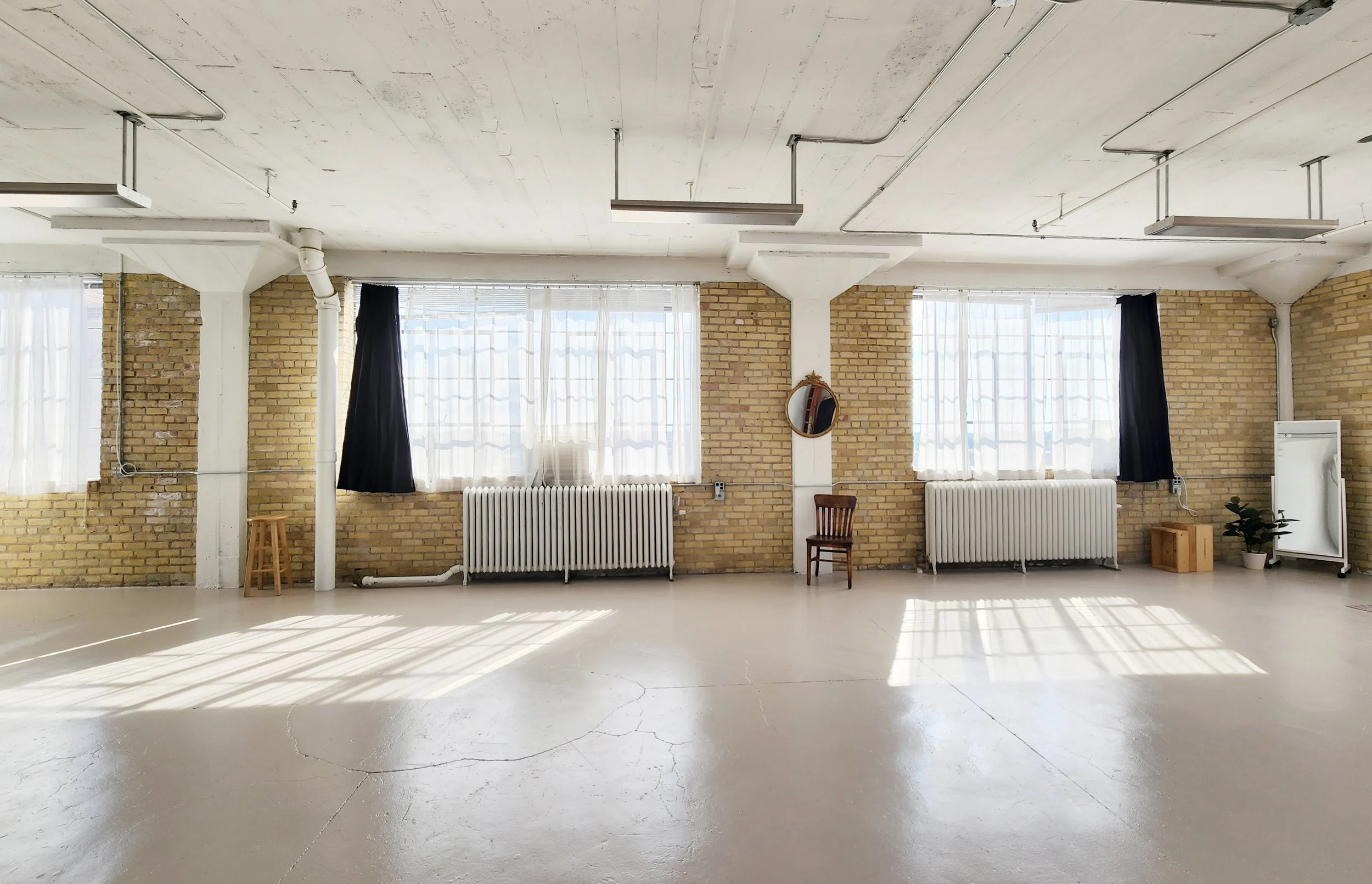 An empty room with large windows, brick walls, white curtains, radiators, and a few chairs and plants.