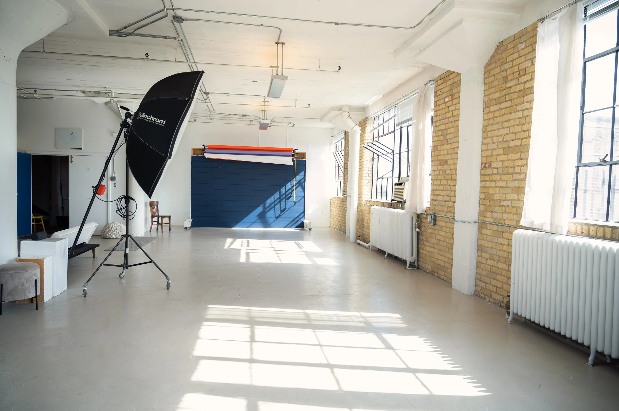 An empty photo studio with large windows and exposed brick walls, featuring photography lighting equipment, a blue backdrop, and natural sunlight streaming in.