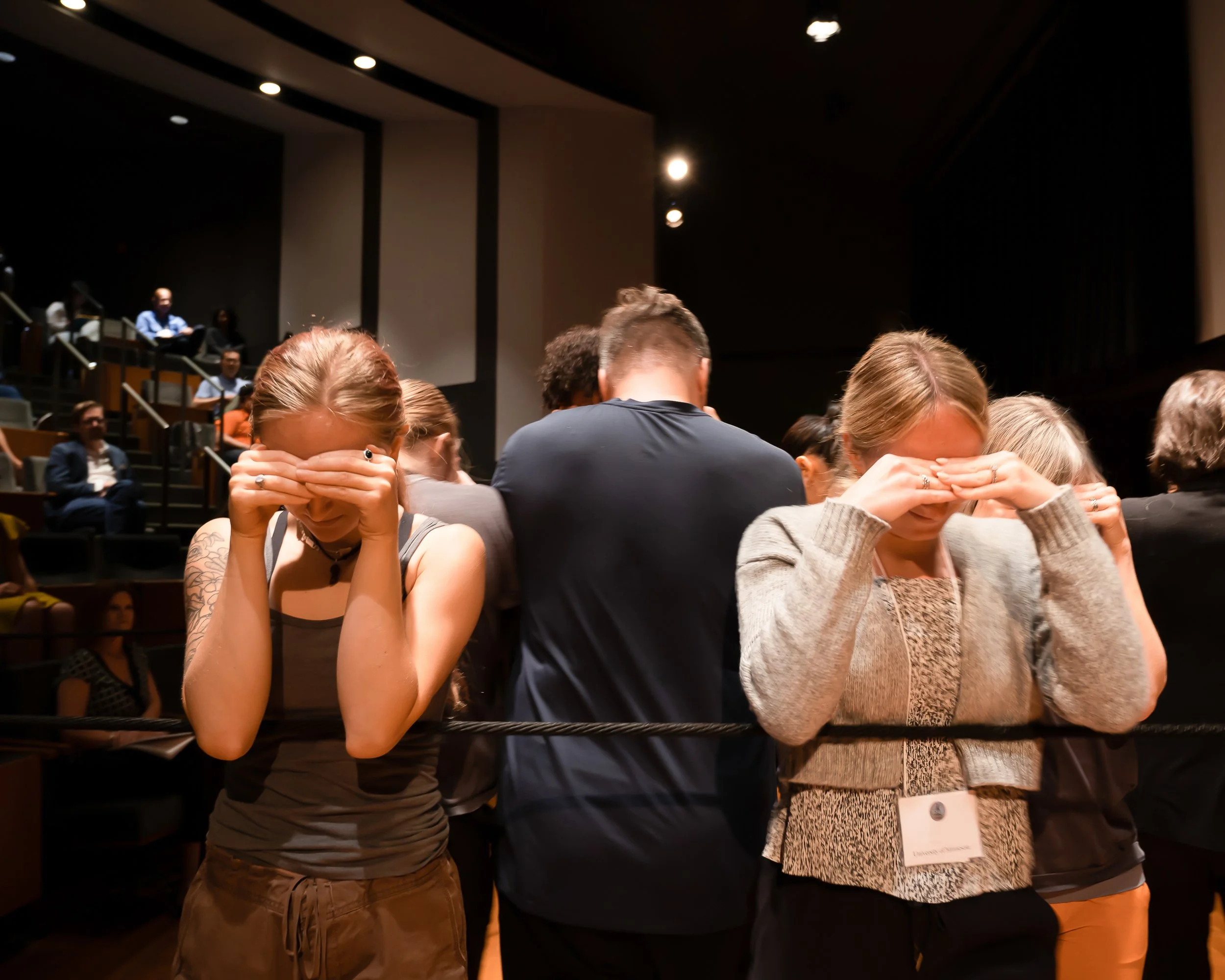 People seated in an auditorium, some appear to be distressed, covering their eyes with their hands. The background shows several individuals, some on a higher level, and the lighting is dim.