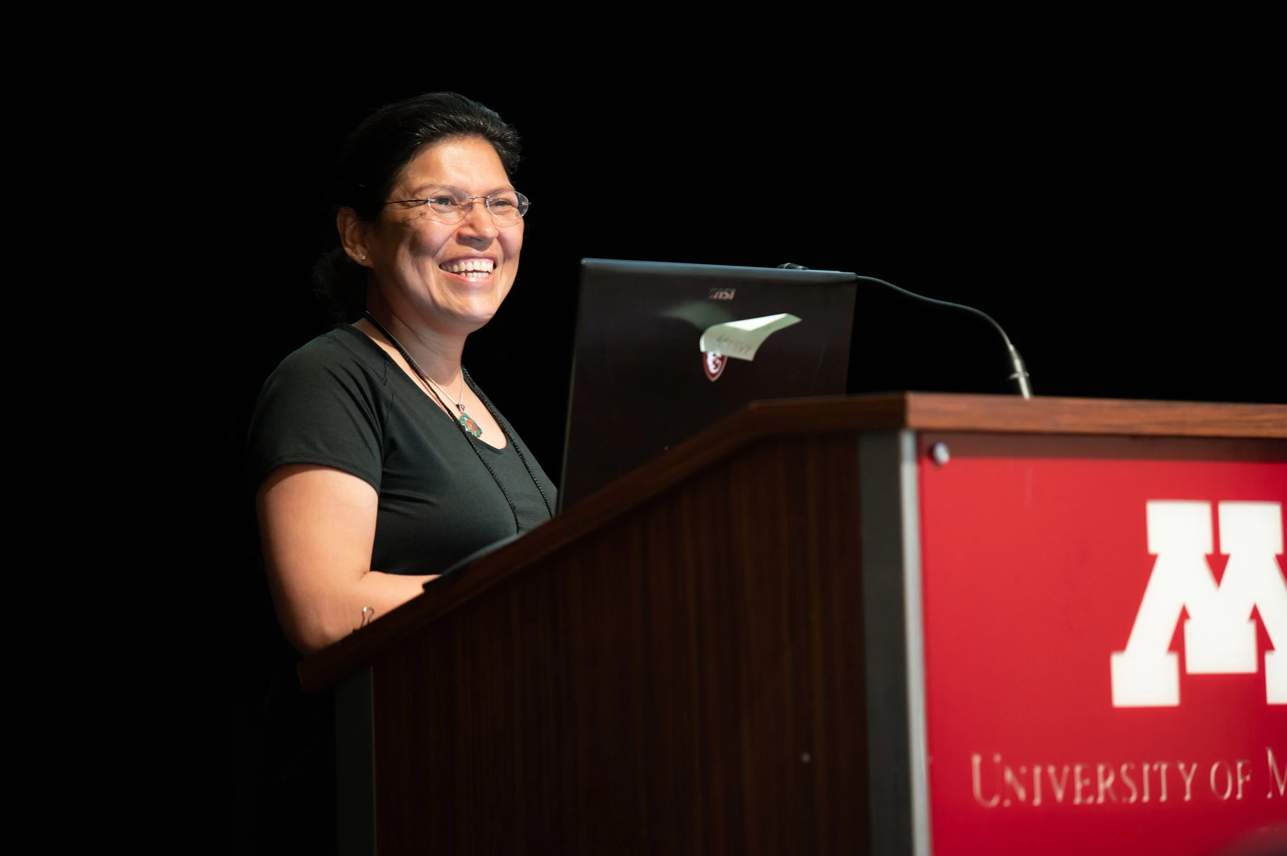 A woman with short black hair and glasses smiling while speaking at a podium with a red sign, in front of a black background.