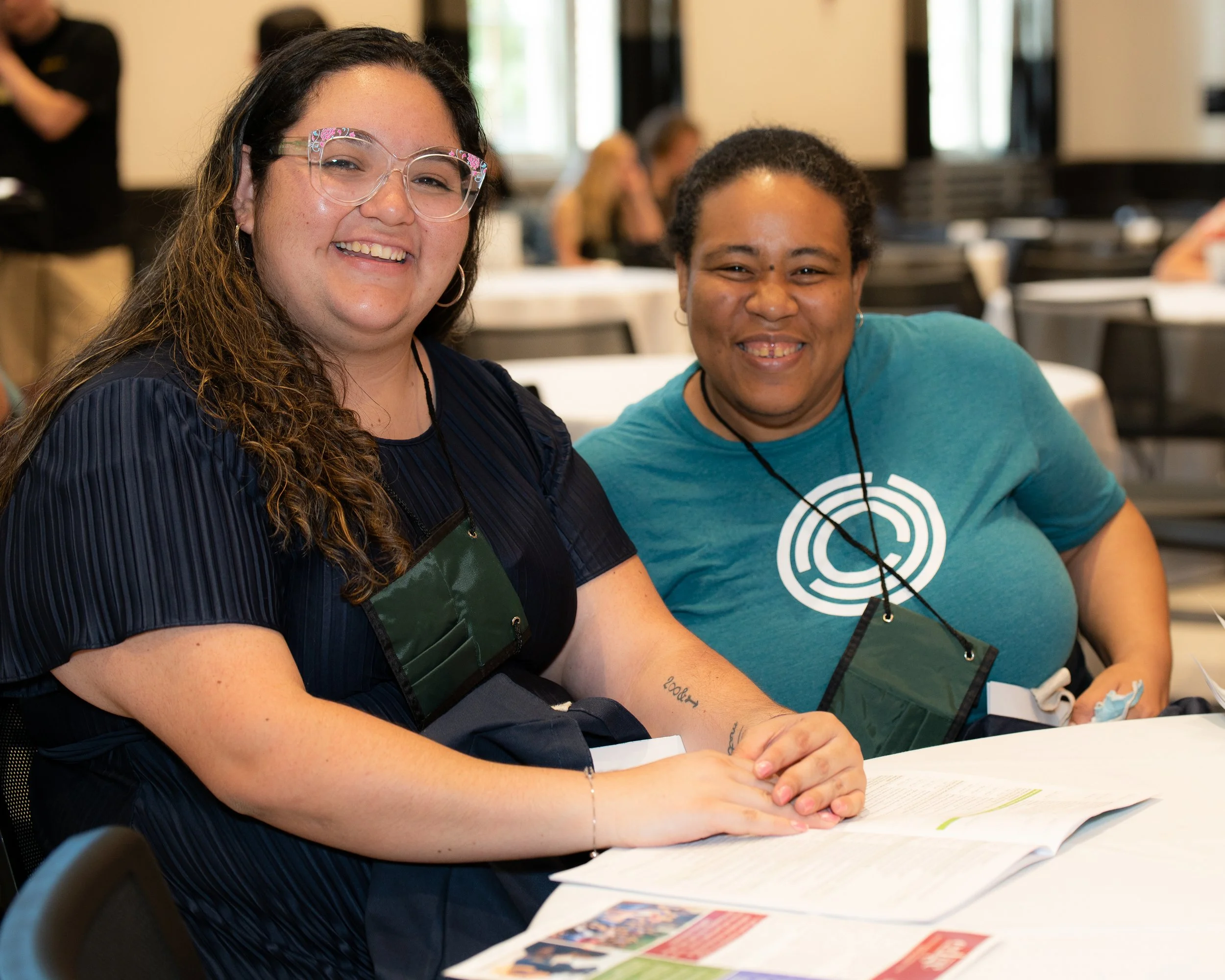 Two women smiling and sitting at a table during an event, with papers and colorful materials in front of them.