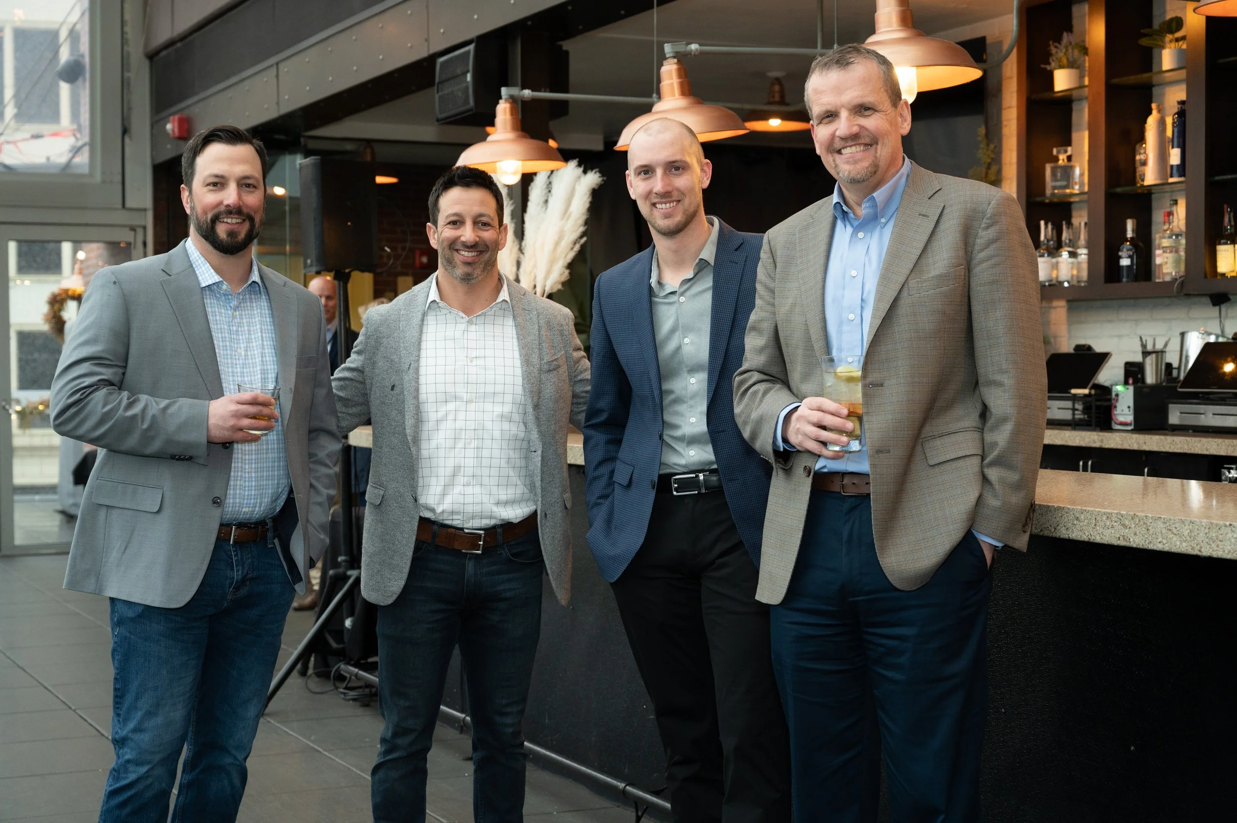 Five men in business attire standing together indoors, smiling at the camera while holding drinks, with a bar and hanging lights in the background.