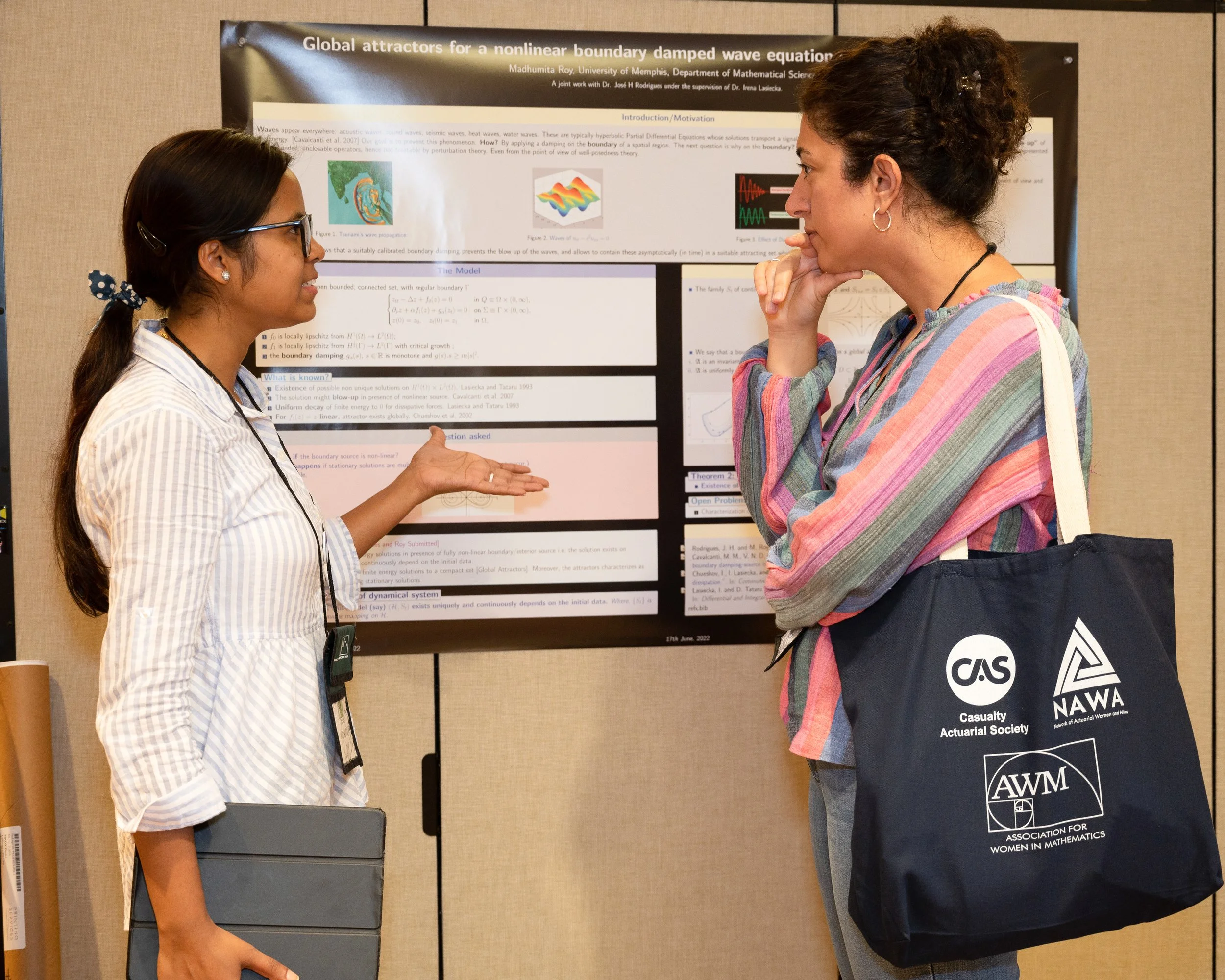Two women engaging in a discussion in front of a scientific research poster about nonlinear boundary damped wave equations. One woman is wearing glasses and a striped shirt, while the other has curly hair and a multicolored striped shirt.