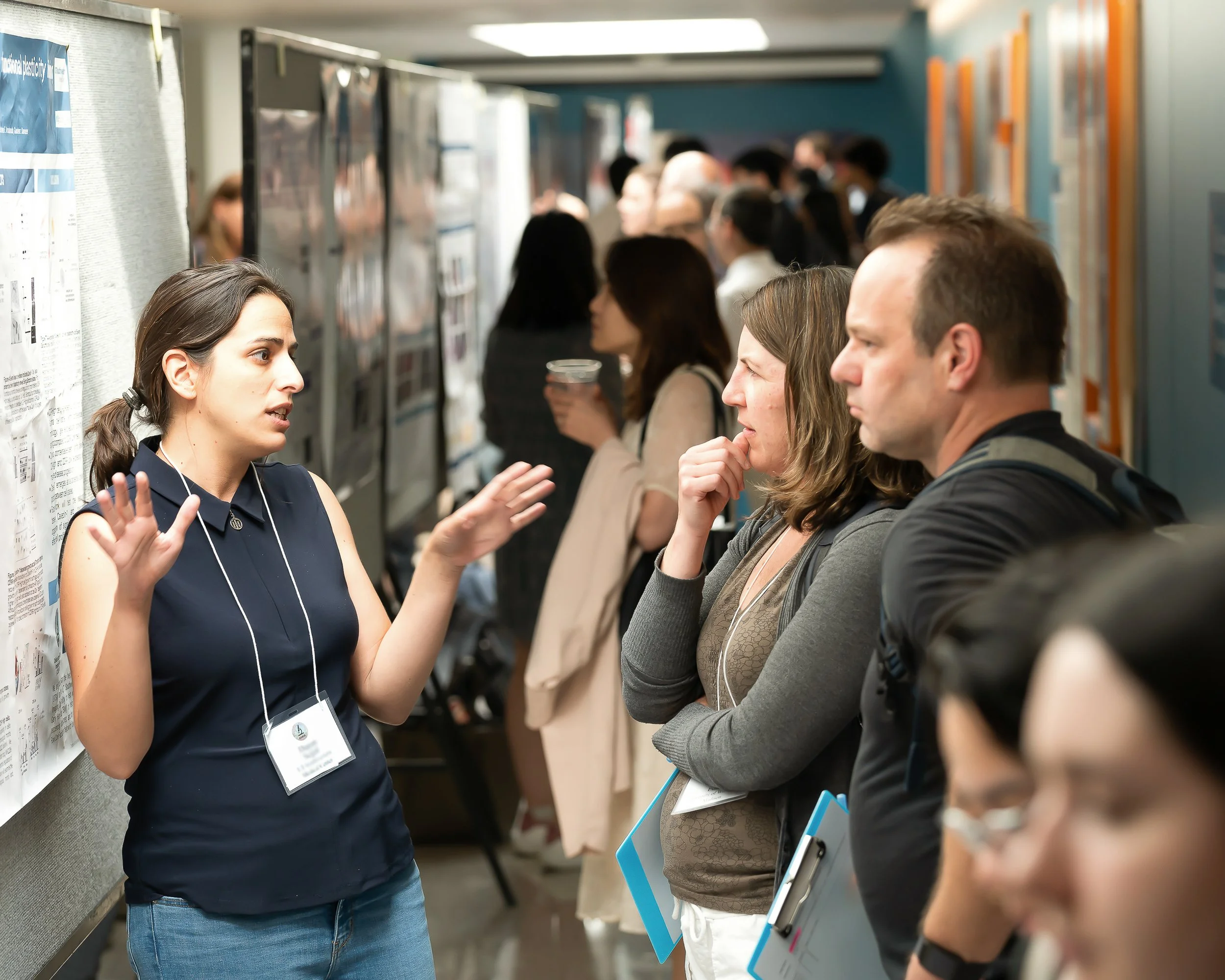 A woman with dark hair in a ponytail, wearing a sleeveless navy top, is explaining information to a line of diverse people in a hallway with posters and lockers, some holding notebooks or cups, during an event or conference.